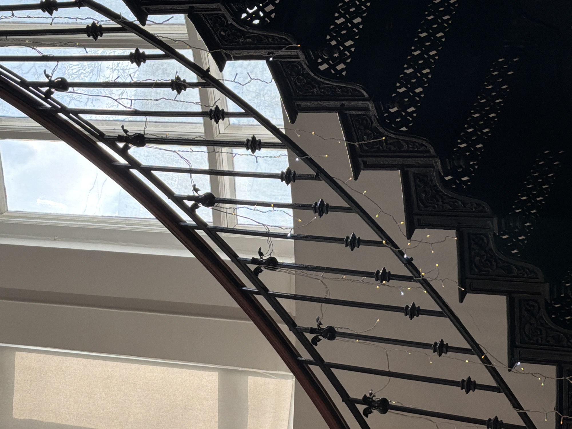 The image depicts a staircase with a decorative wrought iron railing, adorned with small string lights. The staircase is located indoors, with a window visible in the background, offering a view of a cloudy sky. The steps are ornamented with black, decorative trim. The walls are light in color, and a partially visible window shade is in the lower portion of the photo. The overall ambiance suggests an older building with a classic design. The photograph does not depict any people or actions. Moscow, Russia, cannot be confirmed based on the image.