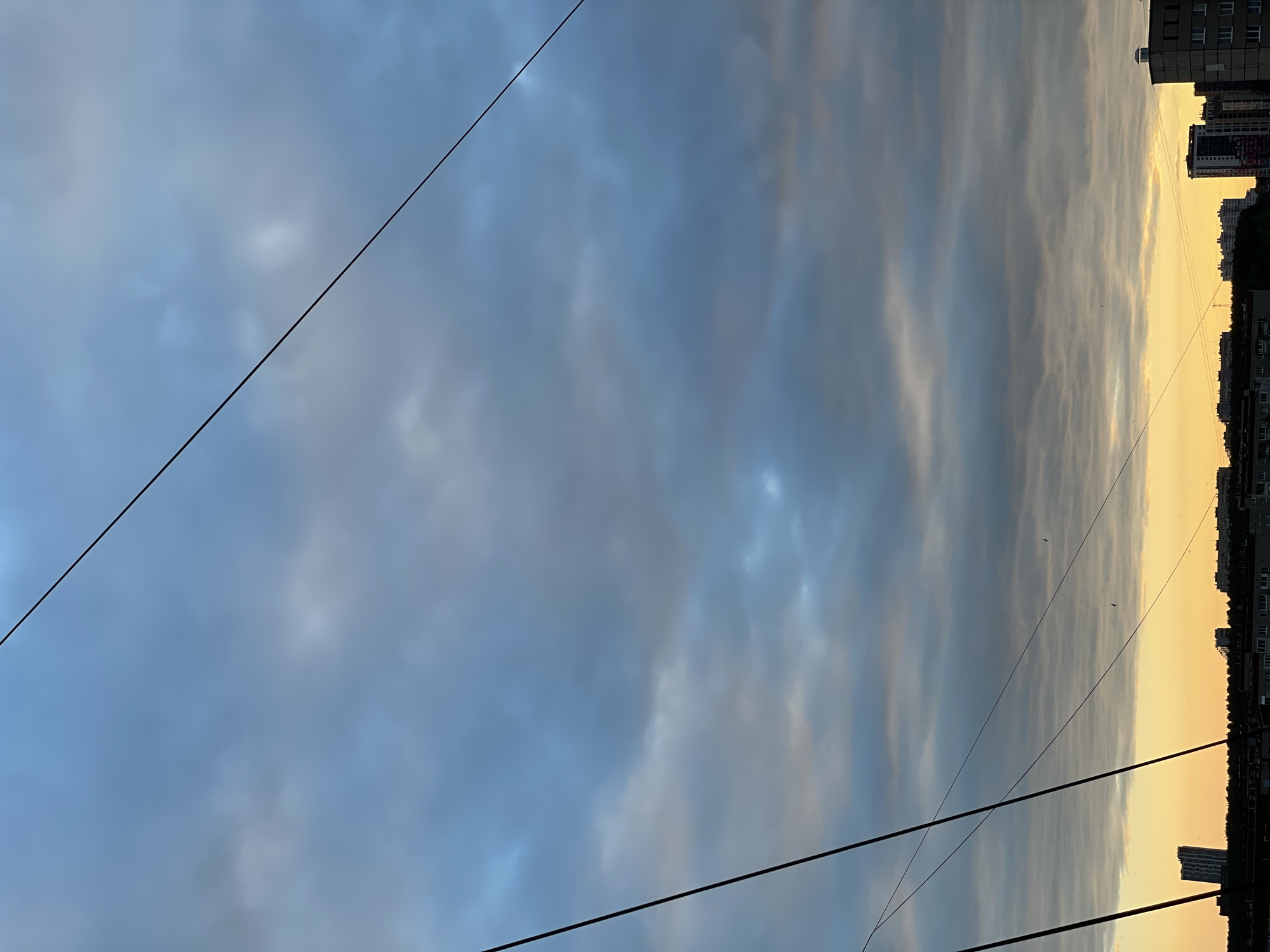 The image depicts a cityscape at dusk, characterized by a dramatic sky filled with varying shades of blue and grey clouds, transitioning to a warm orange and yellow hue near the horizon. The silhouettes of buildings and trees are visible against this sky. Several power lines stretch across the frame, adding a linear element to the composition. In the distance, the outlines of apartment buildings and a taller, modern-looking building can be discerned. The scene is devoid of any visible people or discernible actions, suggesting a quiet, observational moment. The overall impression is one of an urban environment under a transitioning sky, possibly twilight or early dawn, in the city of Izhevsk, Russia.