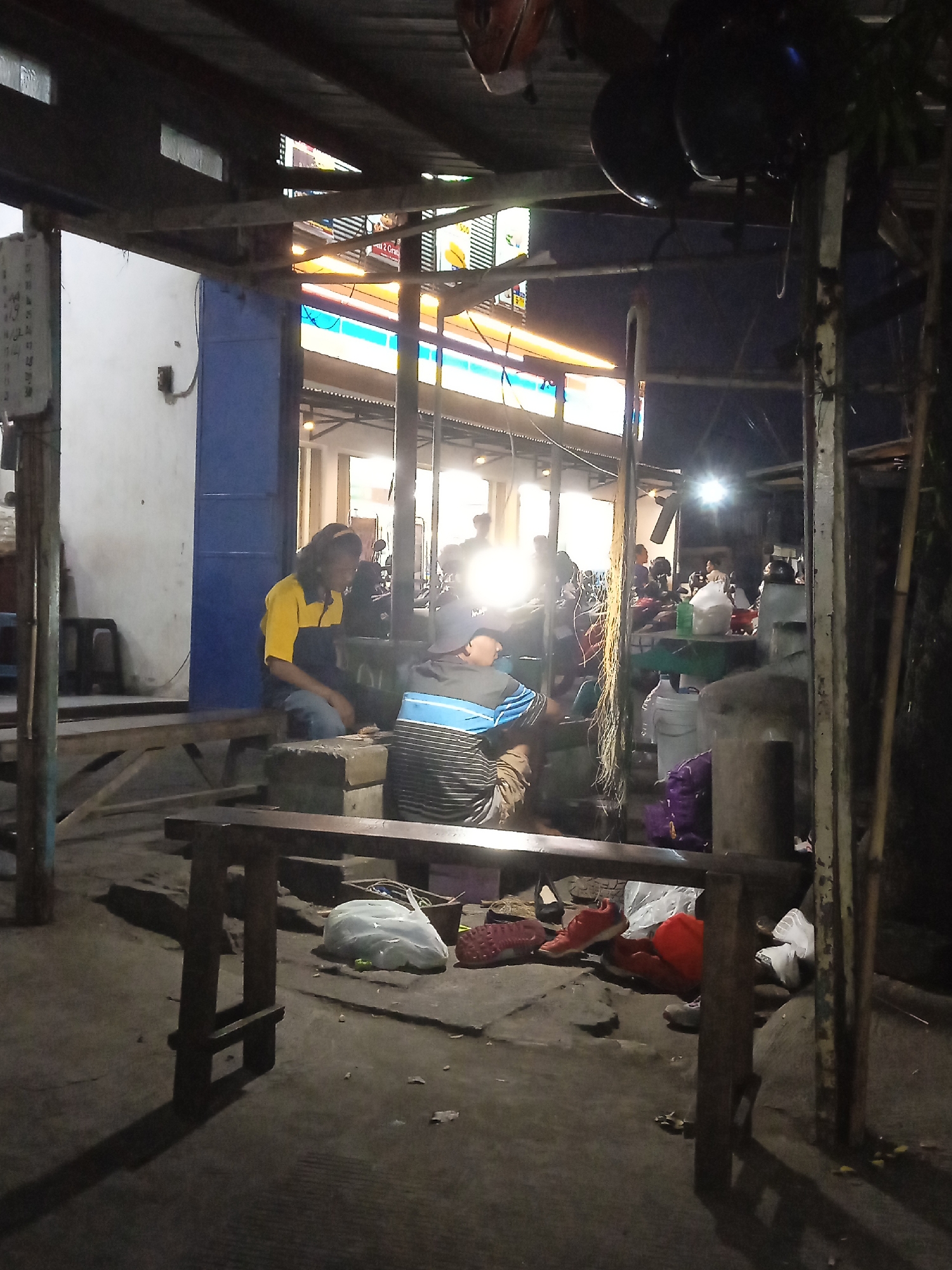 The image depicts an outdoor scene at night, likely a street-side stall or makeshift seating area in Kaliwungu, Indonesia. Two individuals are seated. One, on the left, is a person with long hair wearing a yellow and dark blue shirt and blue jeans. They are sitting on a low wooden bench. To their right, another person, wearing a dark cap and a striped shirt with blue and black bands, is also seated. The background shows a bright illuminated storefront, possibly a convenience store, with neon signs. Various items are scattered on the ground, including plastic bags, shoes (red, orange, and white), and what appear to be large water containers and a purple blanket. The environment is rustic, with visible wooden structures and support beams overhead, and the ground is concrete. A bright light source in the background creates strong shadows.