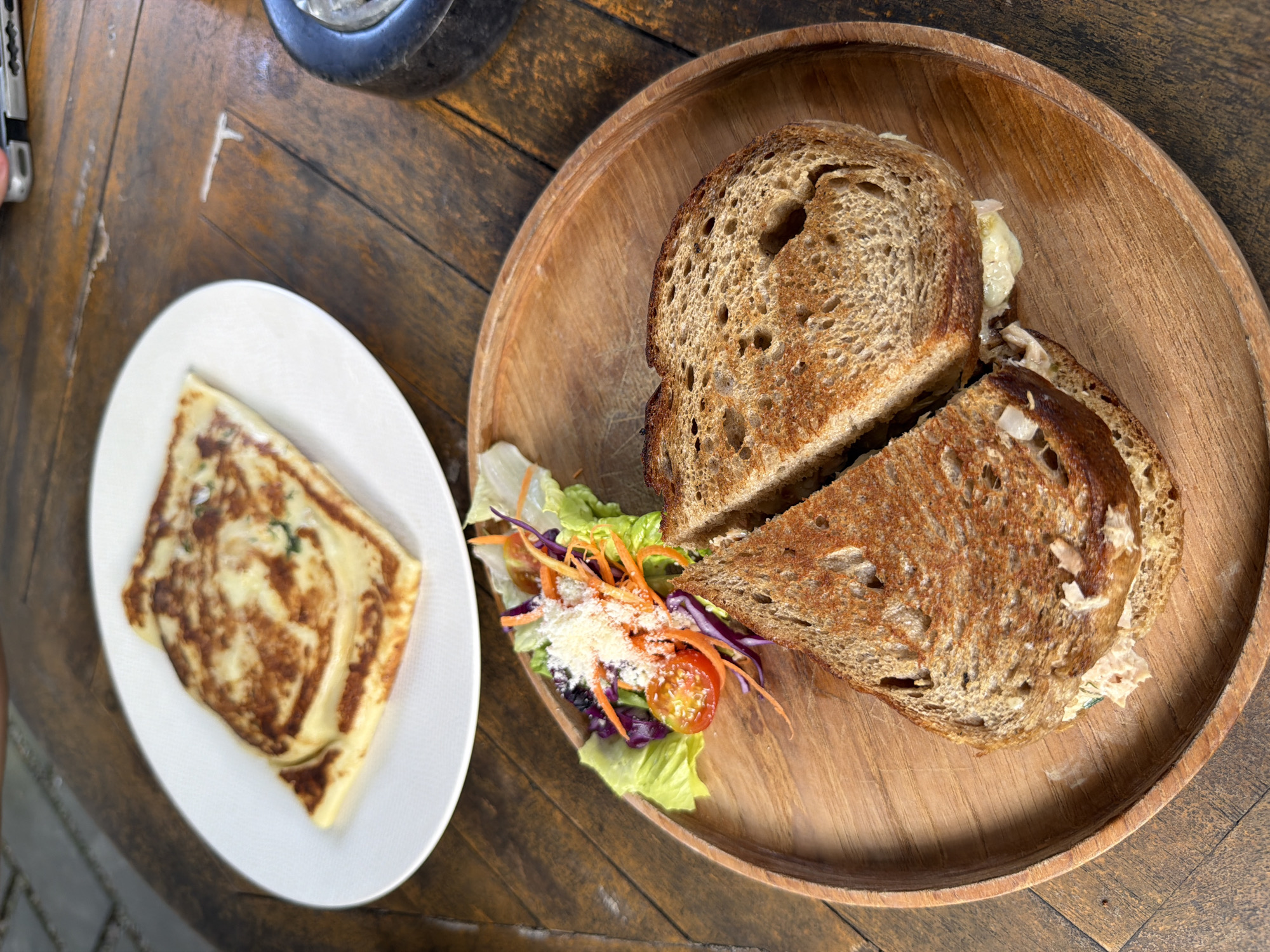 A close-up, overhead shot shows a meal served on a rustic wooden table. The meal consists of a toasted sandwich, cut in half diagonally, served on a round wooden platter. The sandwich is made with whole wheat bread and appears to have a creamy filling. Next to the sandwich, on the same platter, is a small salad with lettuce, shredded carrots, red cabbage, and cherry tomatoes, topped with grated cheese. To the left of the platter, on a white oval plate, is a folded crepe or savory pancake, golden brown with visible green flecks within the filling. The table's wooden surface has a prominent grain and appears to be aged, with some visible wear. The lighting suggests it might be daytime, possibly outdoors given the natural feel of the setting. The scene is focused entirely on the food, implying a casual meal or dining experience. No people are visible in the frame.