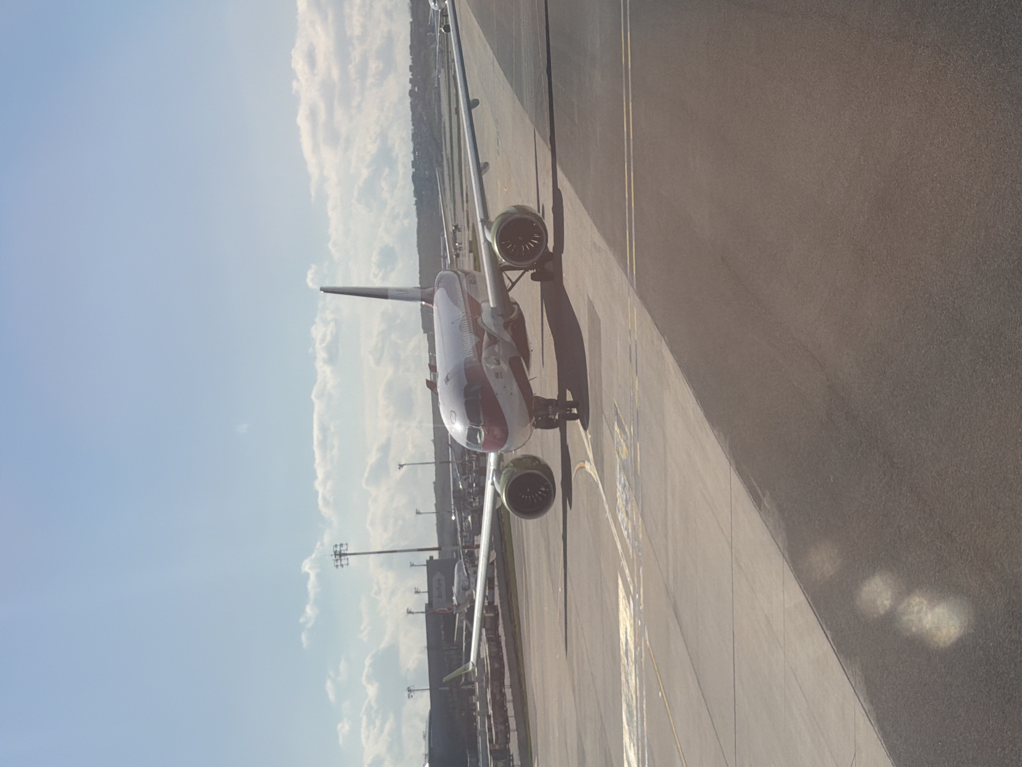 A daytime aerial or elevated view captures a passenger airplane on an airport tarmac. The image is oriented vertically, showing the horizon line across the right side of the frame.
The central subject is a white passenger jet with prominent red markings on its nose cone and extending along the upper forward fuselage. Its left engine is clearly visible beneath the wing, and the landing gear is extended. The aircraft appears to be either stationary or slowly taxiing on the asphalt surface.
The tarmac below the aircraft features various grey and yellow ground markings, including lines and what appear to be numerical or directional indicators, though no specific text is legible. Strong shadows are cast by the aircraft and other structures on the tarmac, indicating bright sunlight.
In the background, beyond the main aircraft, several other airplanes are partially visible, suggesting a busy airport environment. Tall floodlight poles or similar structures are also present. The sky is partly cloudy with substantial areas of clear blue, and the clouds appear white and somewhat puffy. No people are discernible, and no distinct location cues or legible text are visible to identify the airport or airline.