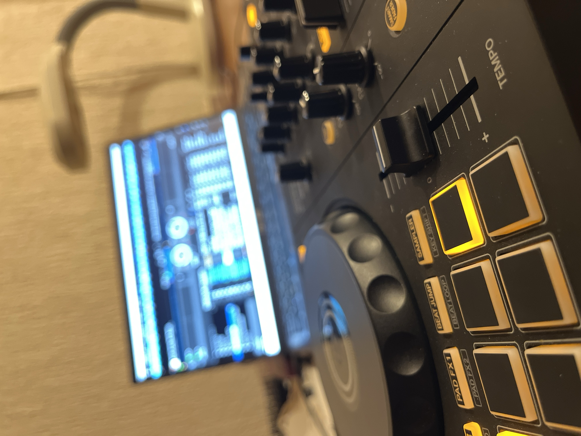 The image depicts a close-up, low-angle shot of a DJ controller and a laptop, likely in a home or studio setting. The DJ controller, in the foreground, is black with various buttons, knobs, and a slider labeled "TEMPO." Several illuminated yellow buttons are visible, including "PAD FX1," "PAD FX2," "BEAT JUMP," "BEATLOOP," and "SAMPLER." A large, dark gray jog wheel dominates the left side of the controller.
Behind the controller, a laptop is open, displaying a screen with what appears to be DJ software, characterized by waveforms and control interfaces rendered in blue and white. The laptop screen is slightly out of focus, as is the background.
In the background, to the right of the laptop, a flexible-armed desk lamp is visible, its head positioned over the workspace. The wall behind the equipment has a textured, light-colored surface, possibly wallpaper. The overall lighting is dim, with the illuminated buttons and screen providing the primary light sources.
There is no indication of the city of Šušanj, Montenegro, in the image. The scene is solely composed of the DJ equipment, laptop, and background elements within an indoor environment. There are no people present in the image.