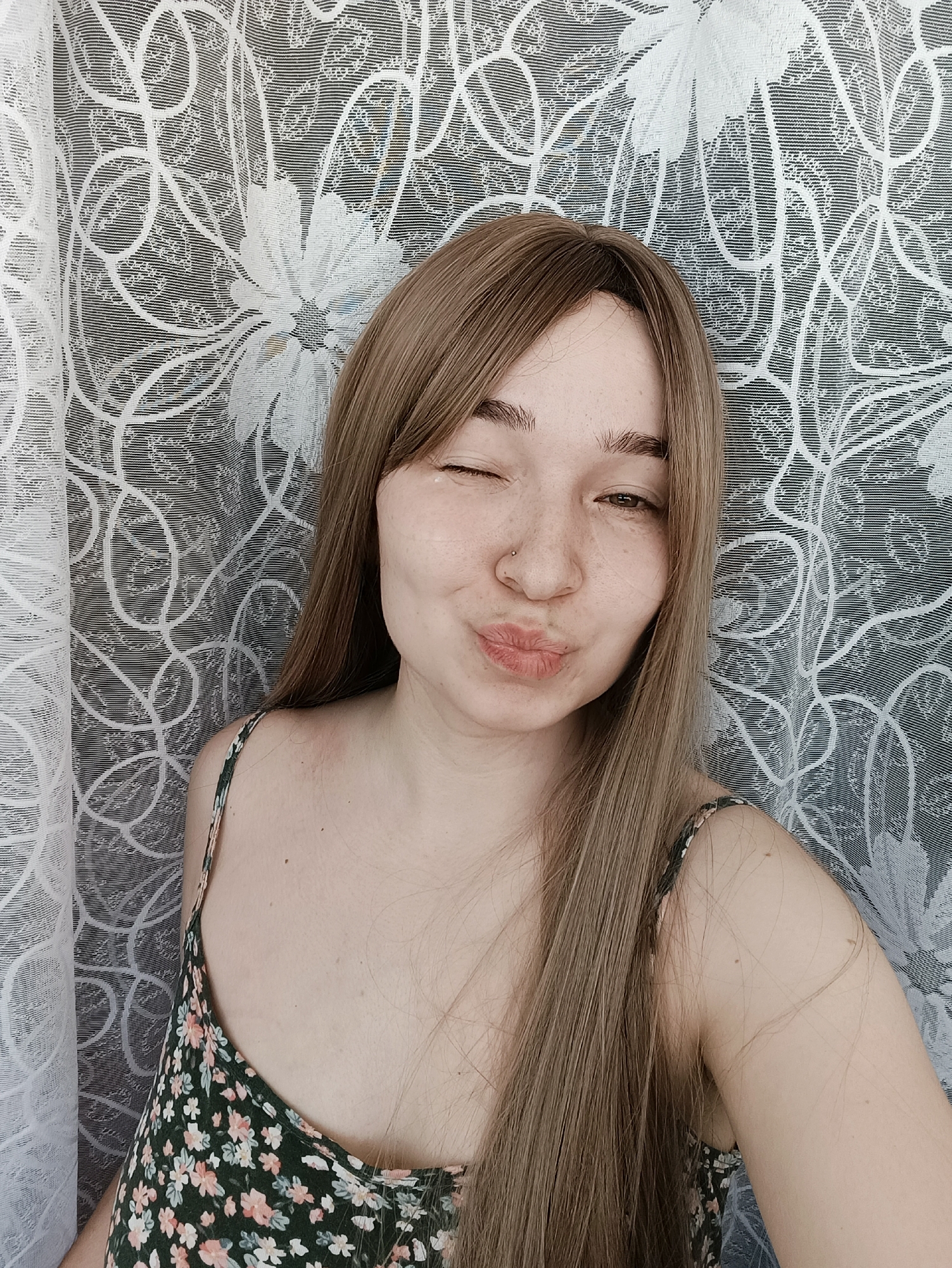 A woman with light brown hair is winking and pouting. She has freckles and a nose piercing. She is wearing a floral-print spaghetti strap tank top. The background is a patterned white and gray curtain. There is no evidence in the image to indicate the location is Izhevsk, Russia.