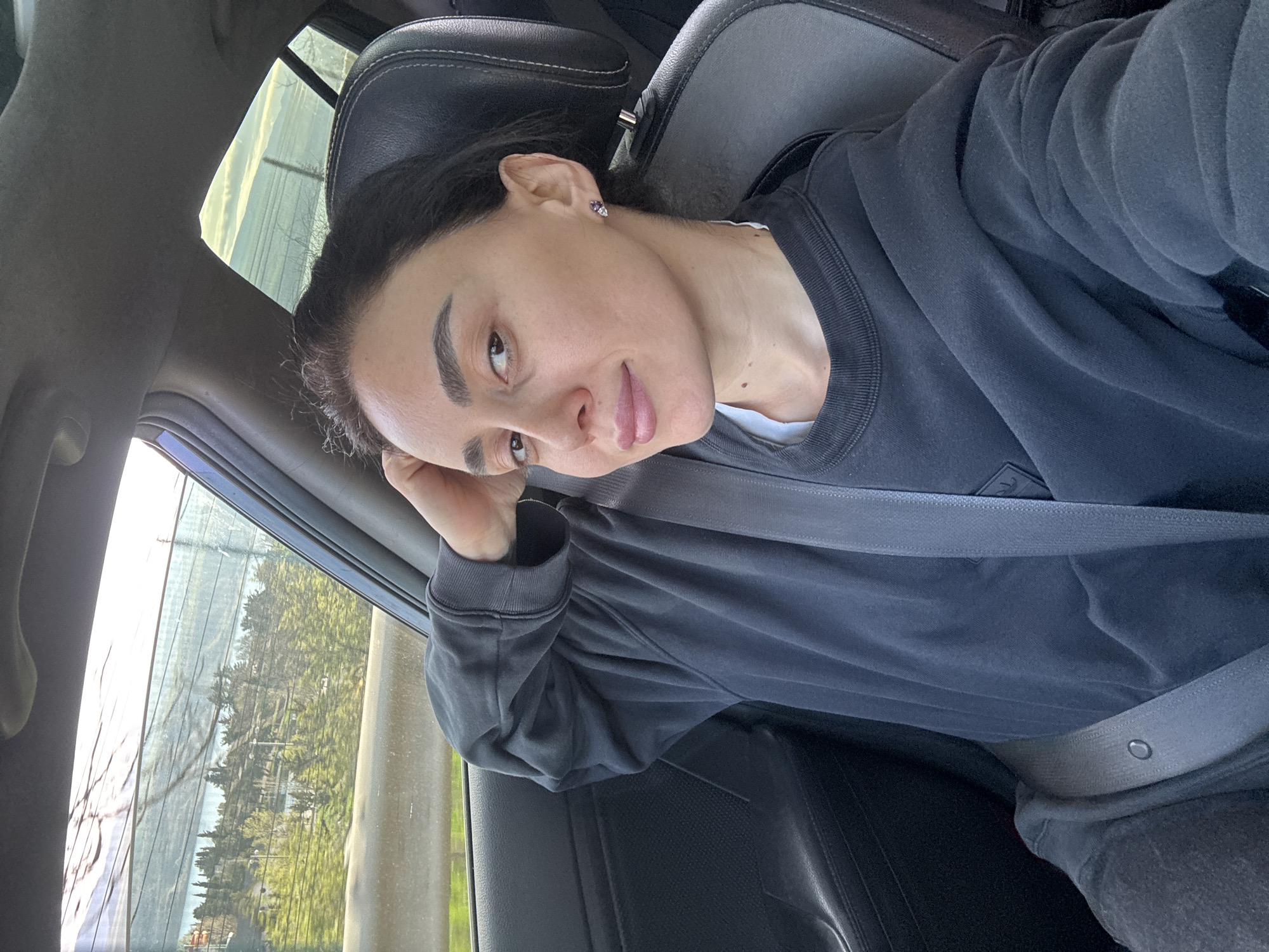 A woman is sitting in a car, wearing a dark-colored sweatshirt and a seatbelt. She is positioned in the passenger seat, leaning her head against her hand. Outside the car window, trees and urban structures are visible. The image suggests she is in a vehicle moving through a city environment. Based on the urban characteristics and tree species, the scene likely takes place in Tbilisi, Georgia.