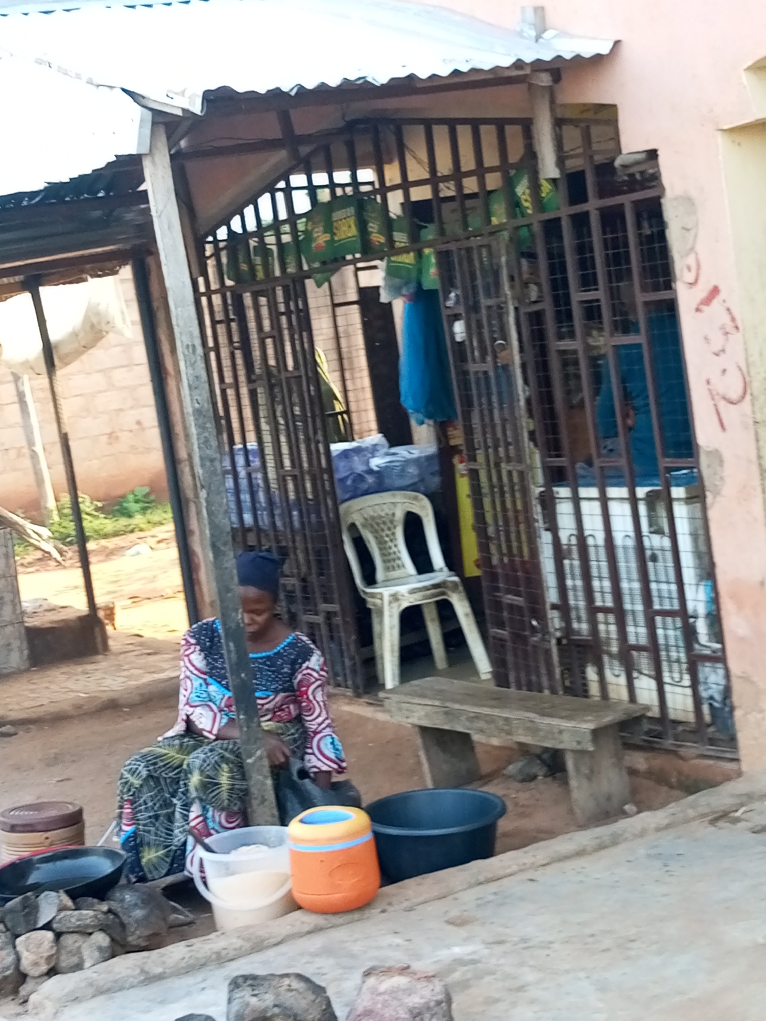 The image depicts a street scene in Gombe, Nigeria, likely in a market or commercial area. In the foreground, a woman wearing a headscarf and a brightly patterned traditional dress is seated near a collection of containers and cooking implements. Among the items are a large black basin, a white bucket filled with a light-colored substance, and an orange and yellow cylindrical container. There are also rocks and a black pan visible. Behind the woman is a simple wooden bench, and beyond that, a structure with a metal grate. Inside the fenced area, a white plastic chair is visible, along with what appears to be a refrigerator. The background shows a building with a corrugated metal roof and brick walls. The overall impression is of a modest, open-air business or household setting.