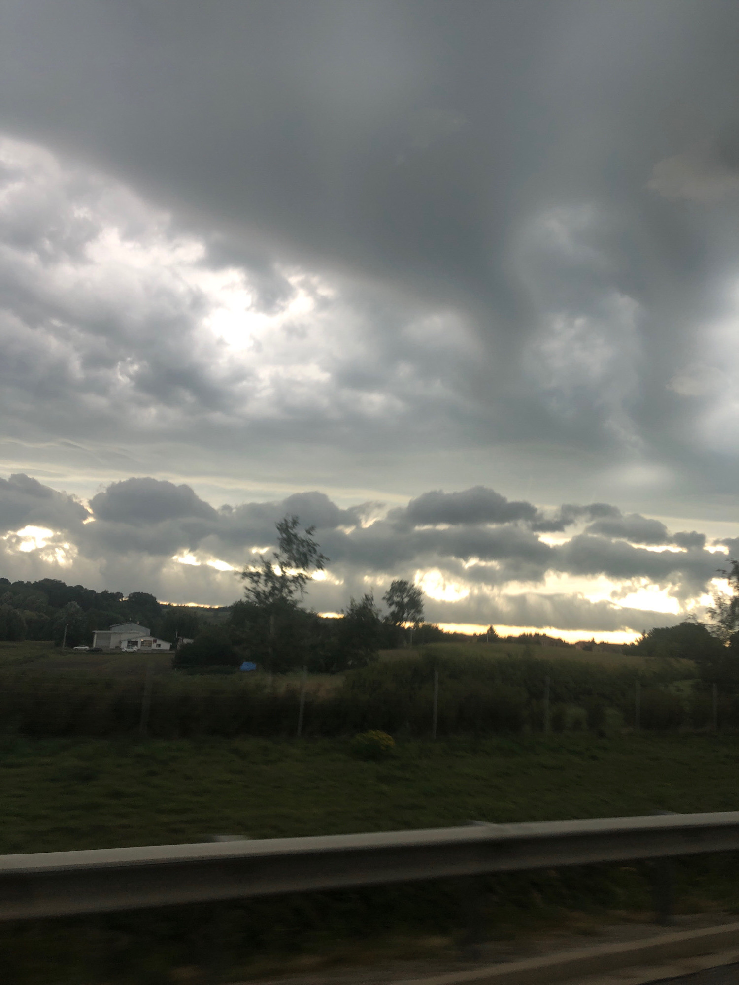 The image depicts a rural landscape under a dramatic, overcast sky. In the mid-ground, a cluster of buildings, possibly a farm or rural housing, is visible. The structures are low-lying and appear to be of a simple design. Surrounding the buildings are fields of grass and trees. The sky is dominated by heavy, dark gray clouds, with patches of brighter light breaking through in horizontal bands, suggesting the sun's rays are piercing the cloud cover. The overall impression is one of a moody, possibly pre-storm or post-storm atmosphere. The location is identified as being in the city of Chłopice, Poland, though the image itself does not contain any specific landmarks to confirm this. A metal guardrail is visible in the foreground, indicating this scene was likely captured from a moving vehicle on a road. There are no people visible in the image.