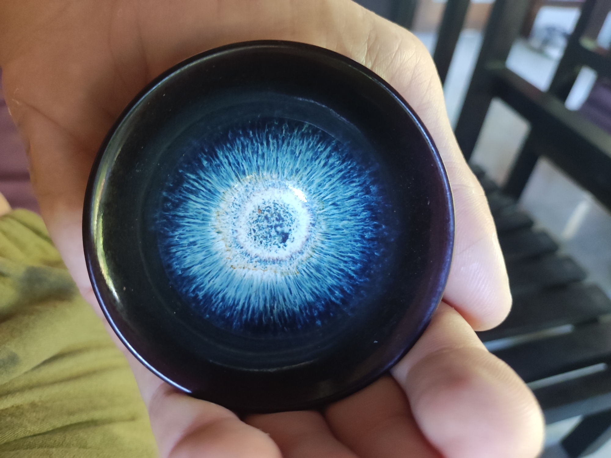 A person is holding a small, dark-colored bowl or saucer in their hands. The interior of the bowl is a swirl of light blue and white radiating from the center, resembling a galaxy or abstract design. The background is partially visible, showing wooden furniture or a structure, and suggesting an outdoor or semi-outdoor setting. Based on the prompt, the location is likely the Ubud District, Indonesia.