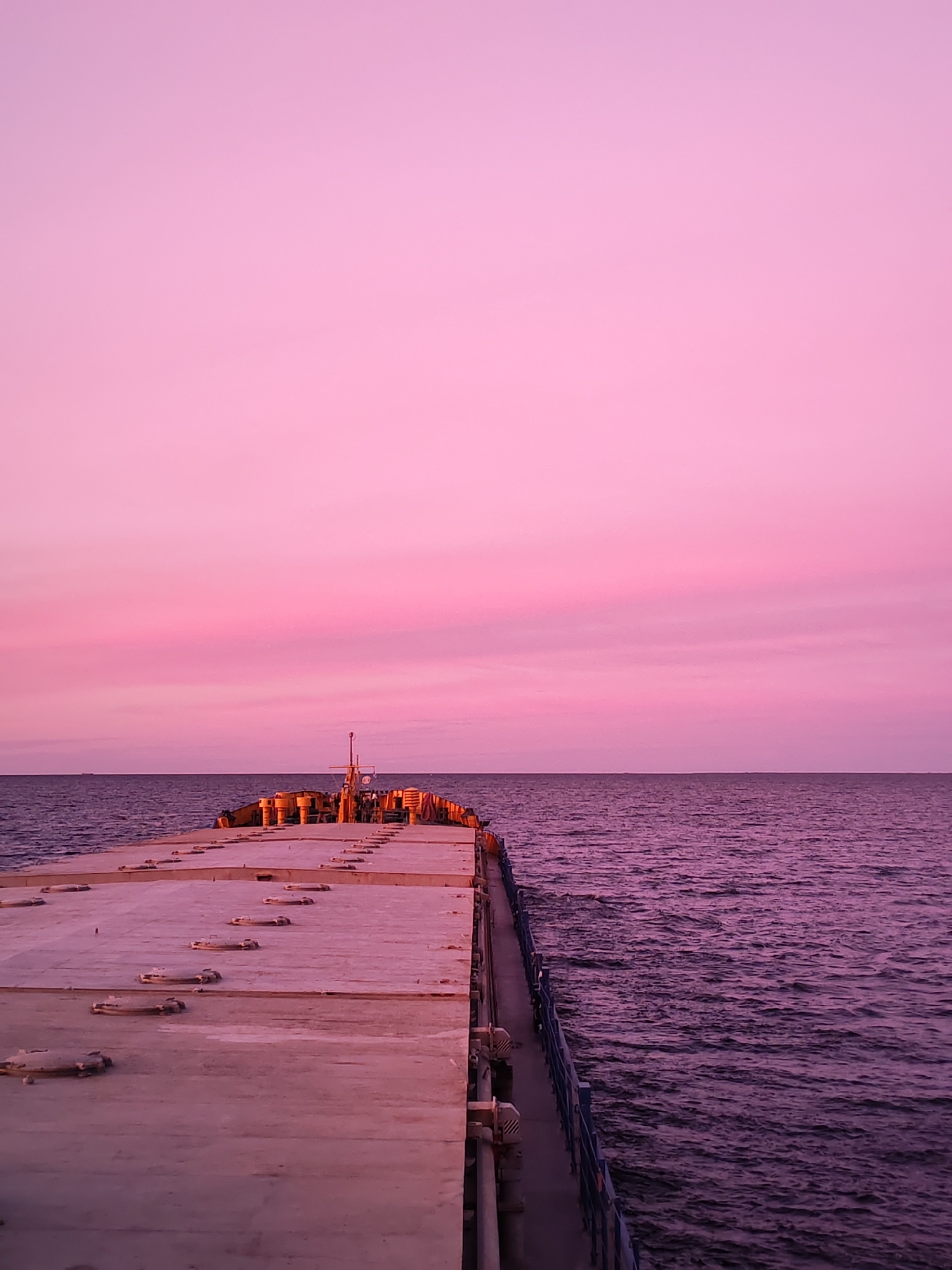 A view of a pier extending into the sea under a pink-hued sky. The pier is a concrete structure with metal hatches. A metal railing runs along the side of the pier. The sea is visible on either side of the pier, with a ripple effect on the water's surface. The sky transitions from a darker pink near the horizon to a lighter shade towards the top of the image. The location is Vasil'evskoe, Russia.