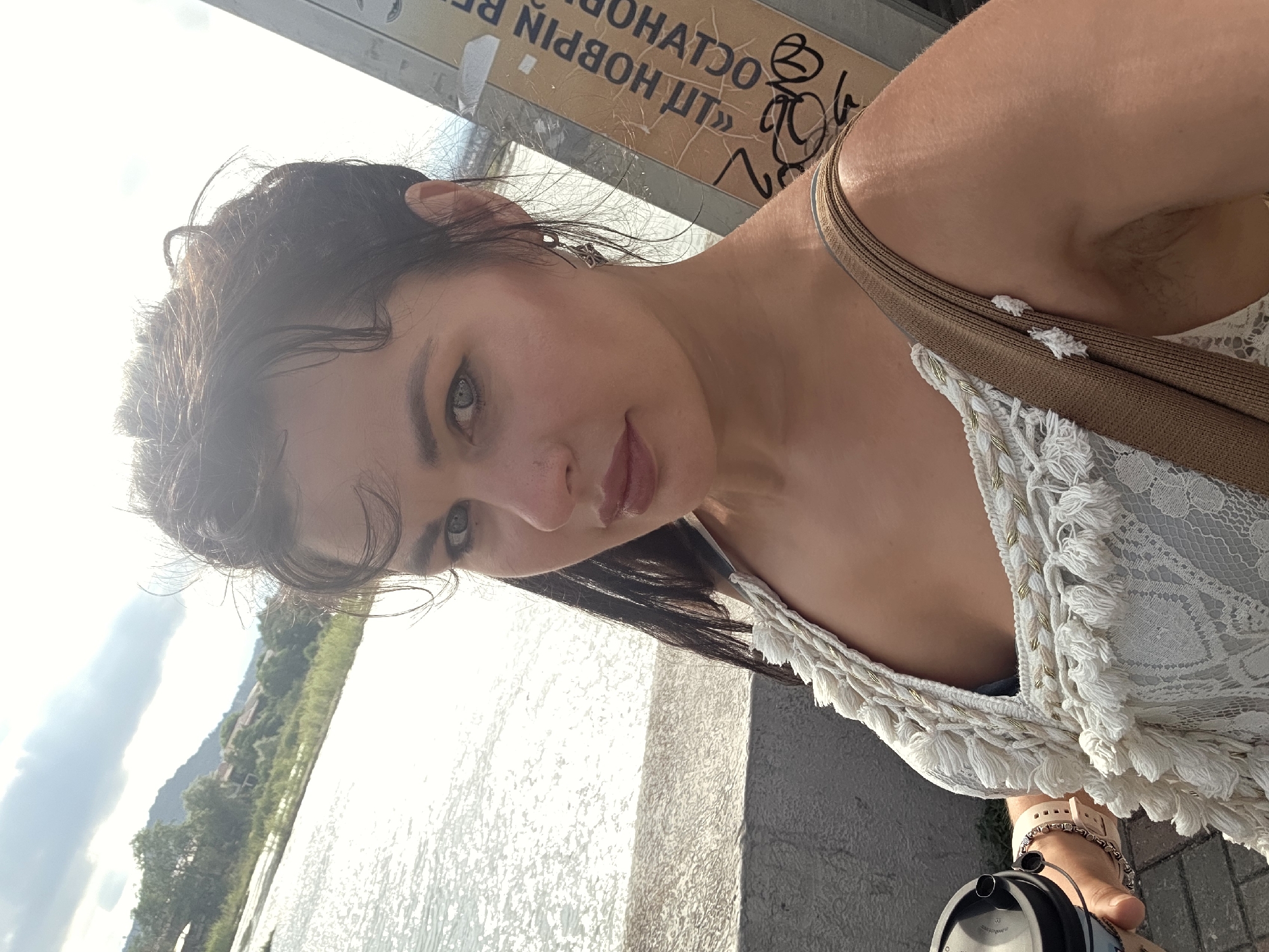 A woman is taking a selfie outdoors. She is wearing a white lacy top with tassels and a brown strap. She has dark hair and is wearing earrings and bracelets. In her left hand, she holds a black cylindrical object. Behind her, there is a body of water reflecting sunlight, and in the distance, there are trees and hills. A sign with Cyrillic writing is visible on the right side of the image. The scene appears to be in Sochi, Russia, likely near a body of water.