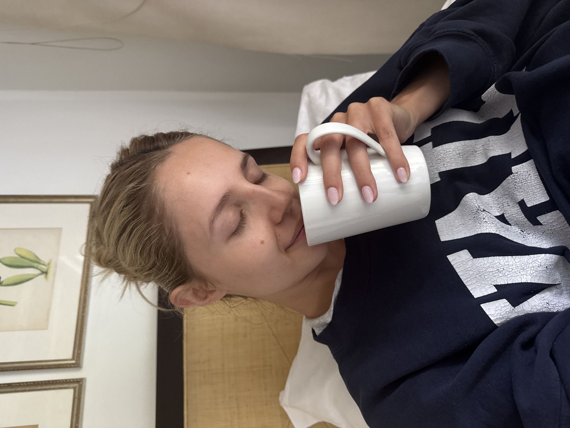 A woman with blonde hair pulled up in a bun is sitting in bed, holding a white mug and drinking from it. She is wearing a dark blue sweatshirt with white lettering. Her eyes are closed, and she appears relaxed. The background shows a headboard, a pillow, and framed artwork on the wall. The setting appears to be a bedroom.