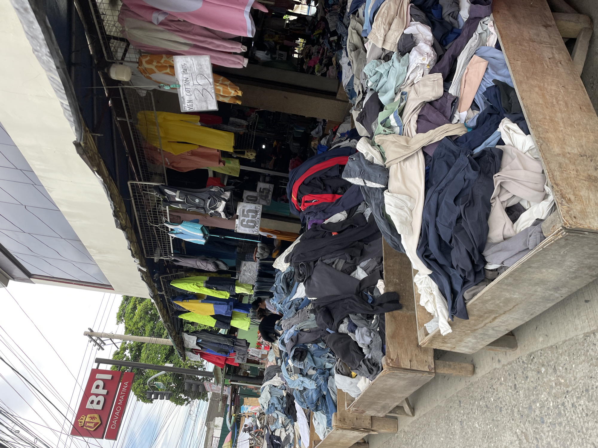 This image depicts an outdoor clothing stall in Davao City, Philippines. In the background, a red BPI (Bank of the Philippine Islands) sign with "DAVAO MATINA" written below it is visible, indicating the location is likely near a branch of this bank in the Matina district of Davao City.
The foreground is dominated by large wooden crates overflowing with piles of second-hand clothing, primarily pants and shirts, arranged for sale. The clothing is sorted by type, with one crate predominantly filled with denim jeans and another with darker colored garments like black pants and shirts.
Above the crates, a covered stall displays more clothing items hanging on racks. These include brightly colored jackets (yellow, blue, red), shirts, and what appear to be children's pajamas or costumes with giraffe and pink patterns. A white sign with handwritten text "MEN COTTON PANTS 30" is hanging in front of some of the clothes. Another sign with "65 JOGGING PANTS 45" is also visible.
The environment is an urban street setting with visible utility wires overhead. A person can be seen in the mid-ground, appearing to be a vendor or customer interacting with the clothing. The overall scene suggests a bustling marketplace environment, common in the Philippines, where affordable second-hand clothing is sold. The pavement and the general appearance of the structures suggest a typical street-side commercial area.