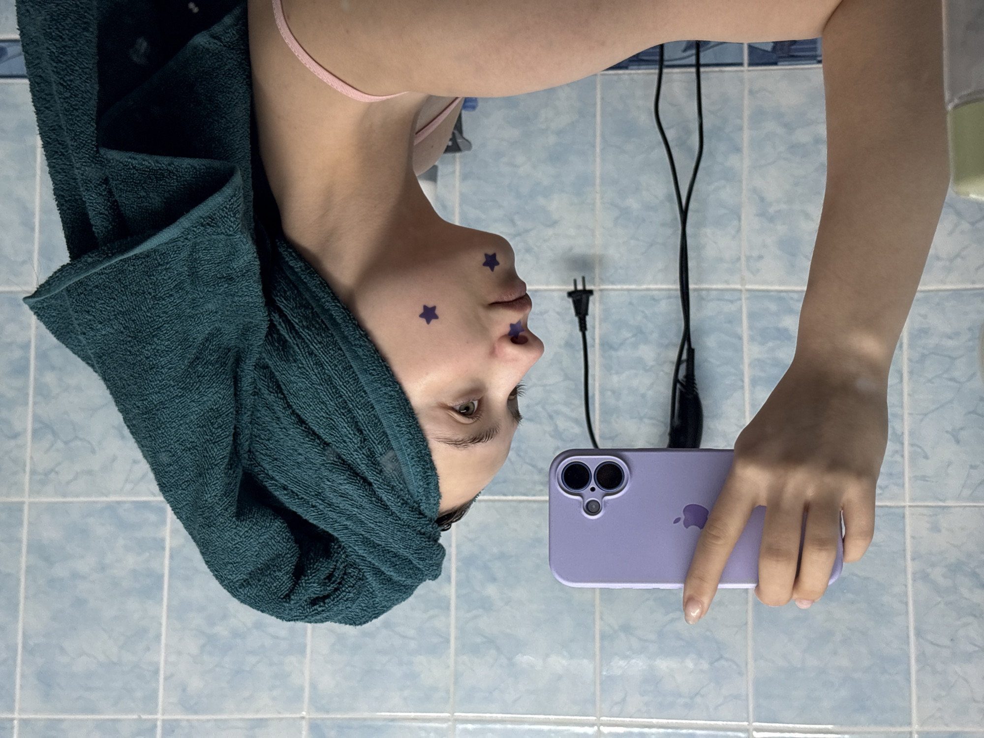 A woman takes a selfie in a bathroom with light blue tiled walls. She has a green towel wrapped around her head. There are three purple star stickers on her face - one on her chin, one on her left cheek, and one above her lip on the right. She holds a purple iPhone in her right hand. A black electrical cord hangs down near the edge of the frame. The image is rotated 180 degrees. The location is Прикамский, Russia.