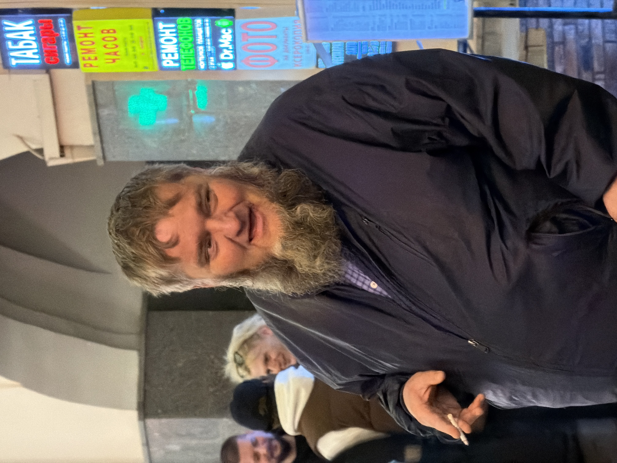 The image shows a man with a gray beard and light brown hair, slightly disheveled, holding a cigarette in his right hand. He is wearing a dark blue jacket over what appears to be a collared shirt. Behind him are other people, including a woman with blonde hair and glasses. In the background, there are signs in Russian, including "ТАБАК СИГАРЫ" (Tobacco Cigars), "РЕМОНТ ЧАСОВ" (Watch Repair), "РЕМОНТ ТЕЛЕФОНОВ" (Phone Repair), and "ФОТО" (Photo), along with a green medical cross. A sign also features "Dr.Mac". The signs suggest this location is in Moscow, Russia.