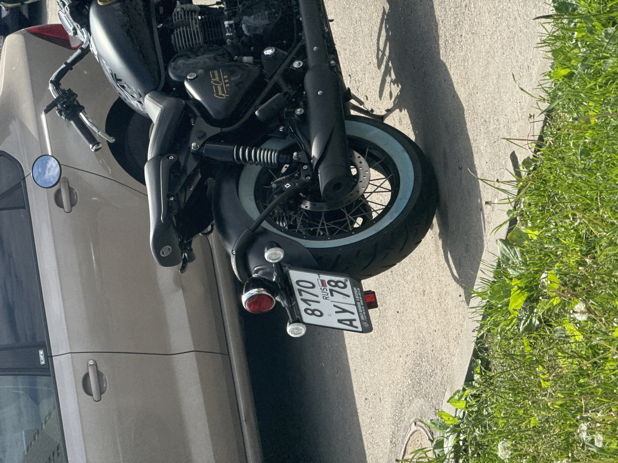 A matte black motorcycle with a classic design is parked on a concrete surface next to a light tan car under bright sunlight. The motorcycle, viewed from its left side, features a prominent "Goan Classic 350" emblem on its engine casing. Its front wheel is equipped with a disc brake, wire spokes, and a distinctive light blue whitewall tire. A circular mirror on the handlebar reflects a cloudy blue sky.
The adjacent light tan car shows part of its side door, handle, and a window with "LIFE" inscribed on its trim.
At the rear of the motorcycle, a Russian license plate is clearly visible, displaying the alphanumeric sequence "8170 RUS AY 78". The license plate holder reads "HARLEY-DAVIDSON С-ПЕТЕРБУРГ", indicating a dealership in Saint Petersburg. A red taillight and two smaller clear lights are mounted on the rear fender.
The setting is outdoors on a sunny day, evidenced by the strong shadows cast on the concrete pavement. Lush green grass borders the concrete surface on the right side of the image.