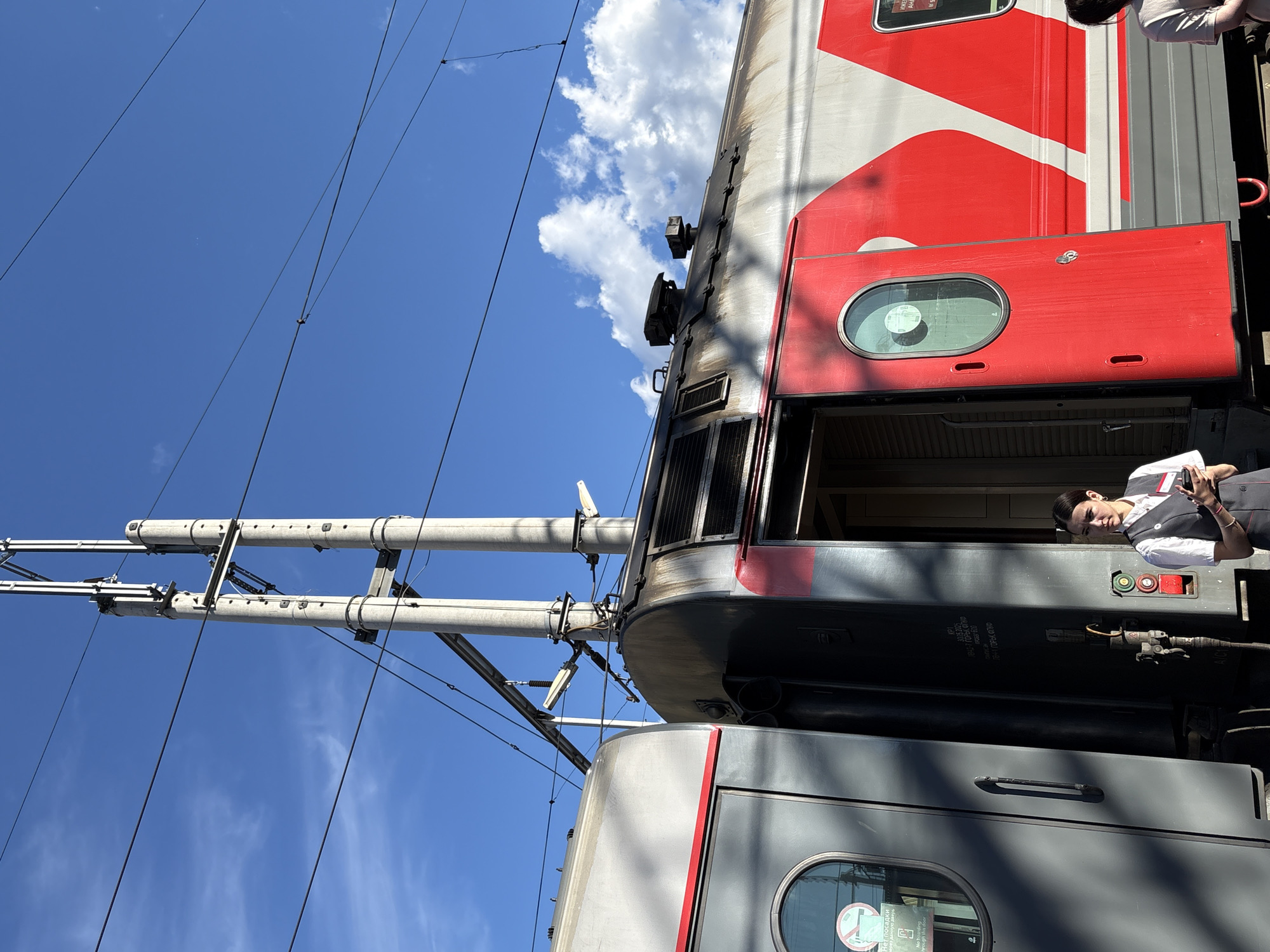The image shows a passenger train in Saratov, Russia. The train is silver and red with the Russian Railways logo. A female employee in a grey uniform is standing near the open doorway, looking towards the camera. Overhead, there are power lines. The sky is blue with some white clouds. Text in Russian is visible on the train's exterior.