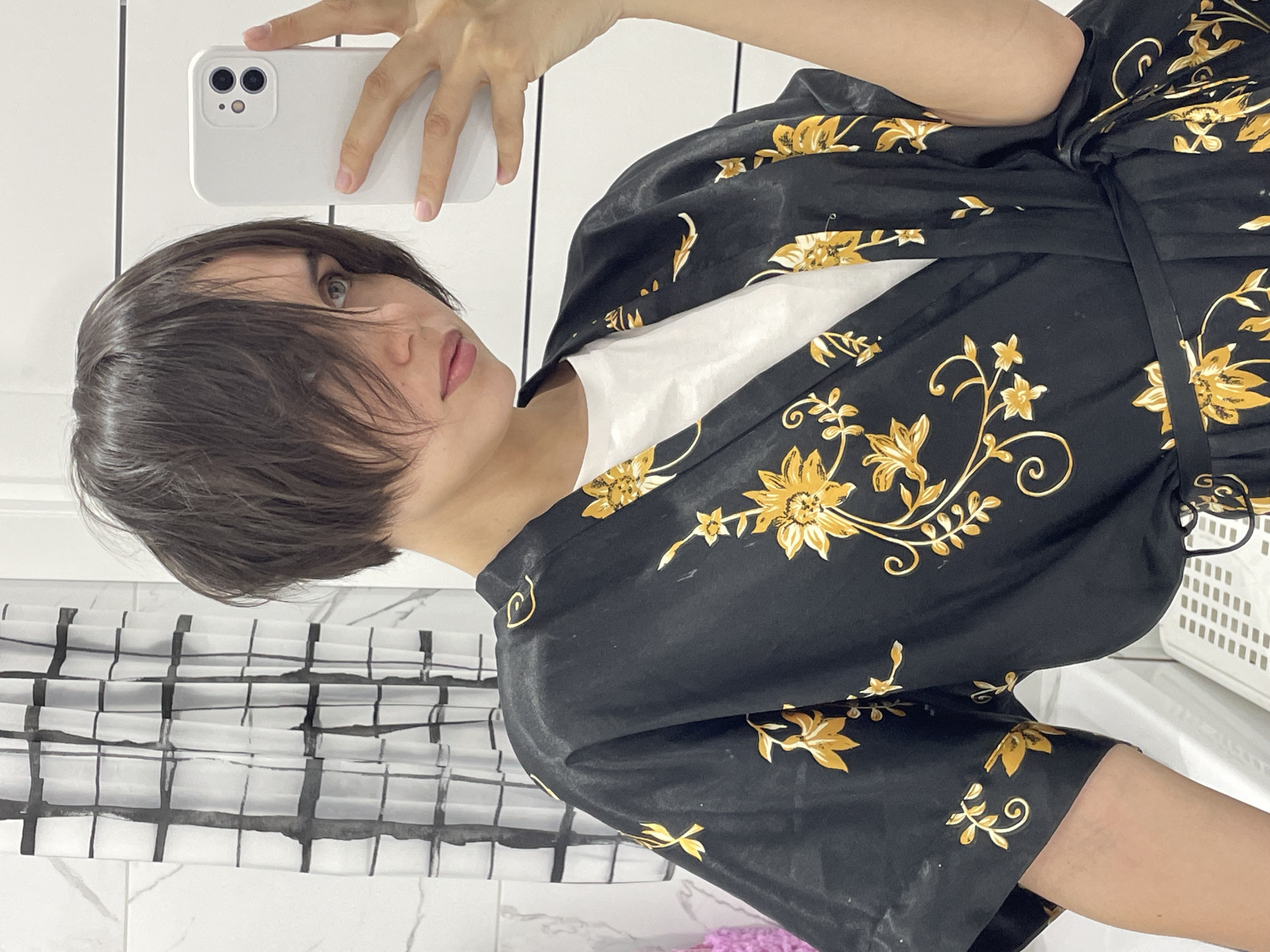 A female individual is depicted in a close-up selfie captured indoors, consistent with a residential or hotel bathroom in Izhevsk, Russia. The person has short, dark brown hair with bangs partially covering the right eye and light-colored eyes, looking slightly to their left. They are wearing a black satin-like robe adorned with a gold floral and vine pattern, tied at the waist, over a white t-shirt. A white smartphone is held in the individual's right hand, positioned to take the photograph. The background elements include white tiled walls, some displaying a subtle grey marble pattern and others featuring horizontal paneling. A white shower curtain with a distinct black grid pattern is visible behind the individual's left shoulder. A purple woven object is partially visible on the far left, and a white plastic slotted container is present in the lower right.