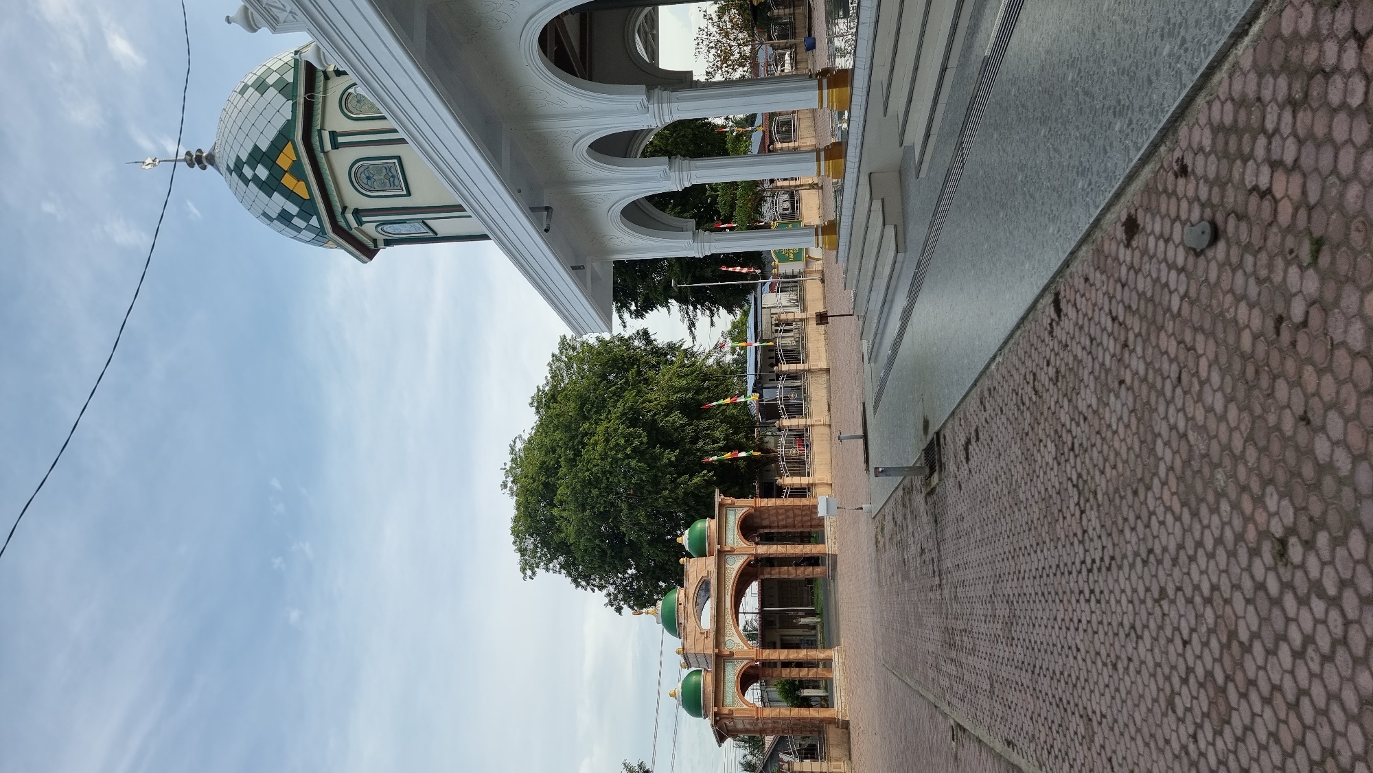 The image depicts an outdoor scene in Meunasahlhok, Indonesia, showcasing a mosque and its surrounding grounds. In the foreground, a patterned red brick pathway leads towards a modern, white-pillared structure with arched openings. To the left, an ornate, arched gateway with green domes stands as an entrance. A large, leafy green tree is visible behind the gateway. The sky is partly cloudy with a few scattered white clouds. Several colorful flags on poles are planted in the ground between the gateway and a fence. The overall atmosphere suggests a tranquil, possibly early morning or late afternoon setting. There are no people visible in the image.