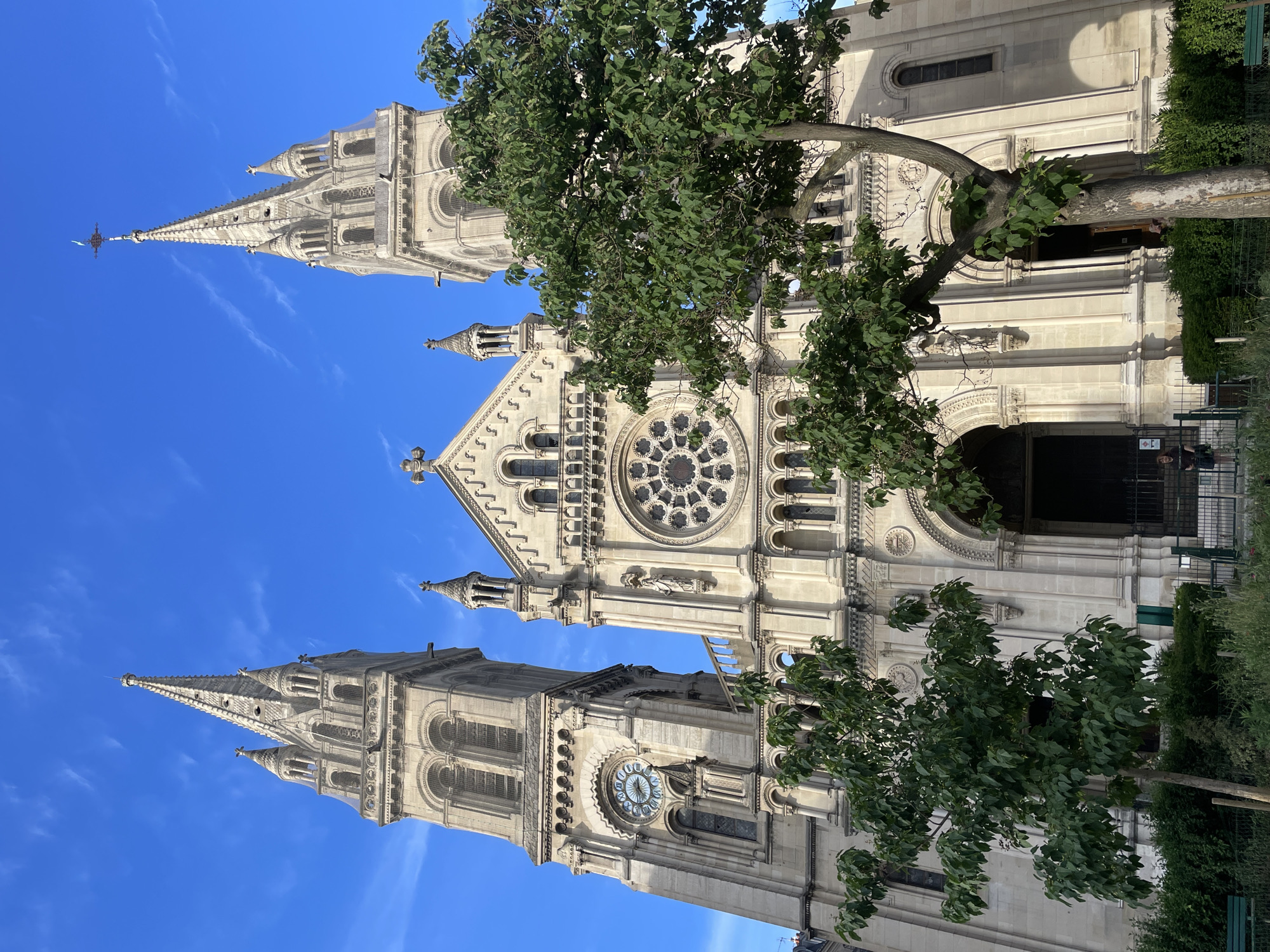 The image displays a grand, ornate stone church, the Eglise Saint-Augustin, prominently featured against a vibrant, clear blue sky dotted with scattered wispy white clouds. The original image and its cropped hints are all presented rotated 90 degrees clockwise; when viewed upright, the church rises majestically.
The architecture is a blend of neo-Romanesque and neo-Byzantine styles, characterized by light-colored stone and intricate detailing. The church features two tall, slender spires, each adorned with finials and topped by crosses. The left spire has a distinctive weathercock or similar decorative element at its very top. Between the towers, the main facade showcases a large, ornate rose window with detailed circular tracery. Below the rose window, a series of arched entrances are visible. The right tower distinctly features a large, round clock face with Roman numerals. The entire facade is rich with architectural elements including numerous arched windows, decorative columns, intricate carvings, and statues positioned in niches.
Two large trees with dense green foliage partially obscure the church's facade and spires, one on the left and another on the right, adding a natural contrast to the stone structure. In the lower parts of the image, near the church's base, glimpses of green fencing and shrubbery can be seen.
The scene depicts a bright, sunny daytime, evidenced by the clear sky and well-defined shadows cast by the church and trees. No specific activities are taking place, and aside from the Roman numerals on the clock face, no other discernible text is visible. The imposing scale and detailed craftsmanship of the church are the focal points, set within an urban environment in Paris, France.