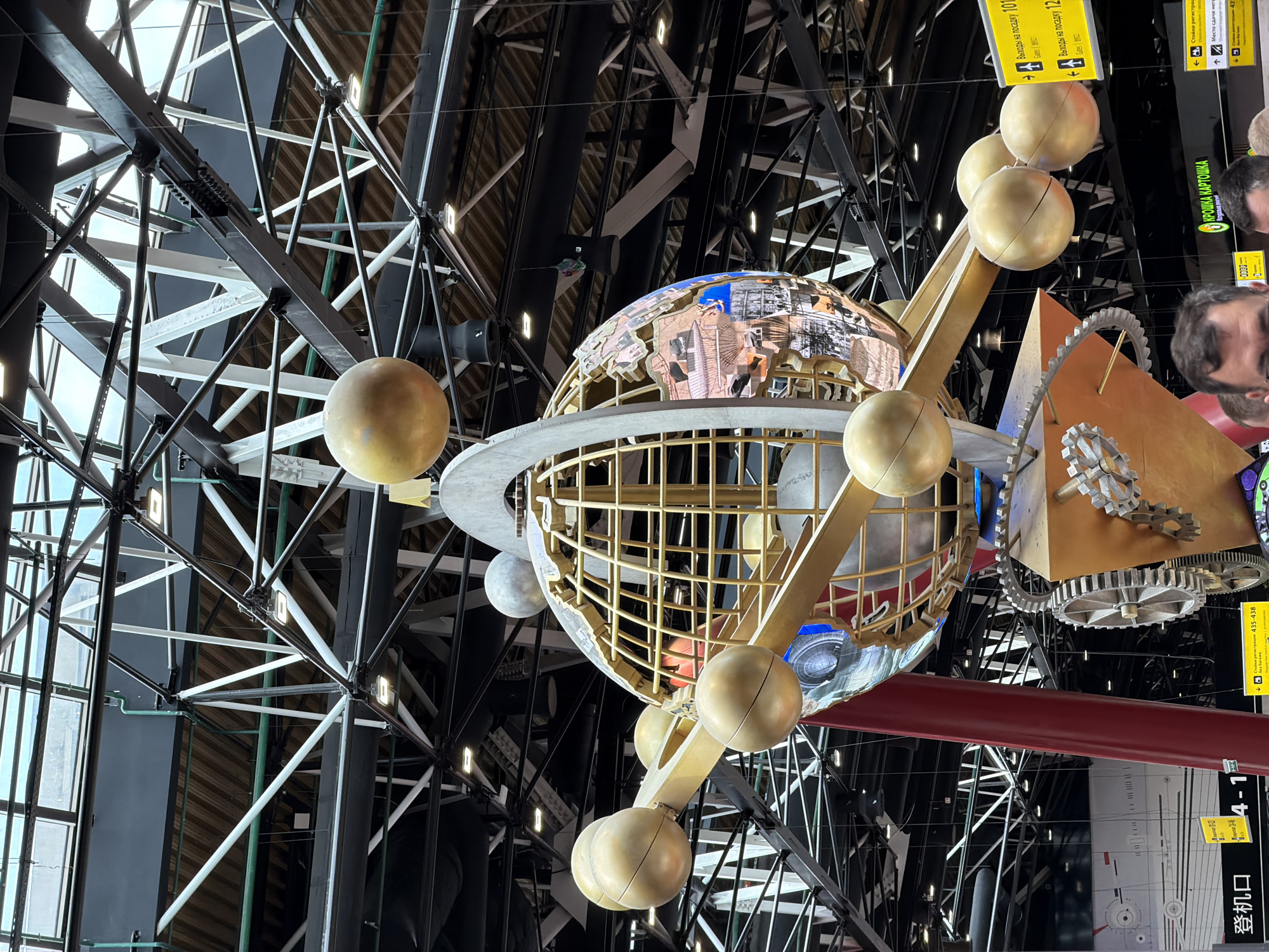 The image shows a large, complex mobile suspended from the ceiling of what appears to be a transportation hub, likely an airport or train station. The mobile includes a metallic globe with a grid structure, adorned with golden spheres and additional planetary elements. Adjacent to the globe is a stylized gear-and-pyramid structure.
The mobile is suspended against a backdrop of a complex network of black metal beams and supports typical of large modern architectural structures. The lighting is a mix of natural light coming through high windows and artificial lighting from small fixtures attached to the beams. Signage with text in Cyrillic and other languages, including departure gate information, is visible. The lower right corner shows part of a person's head, suggesting the size and scale of the artwork.
Based on the presence of Cyrillic text and recognizable signage, the mobile is probably located in a transportation hub in Khimki, Russia.