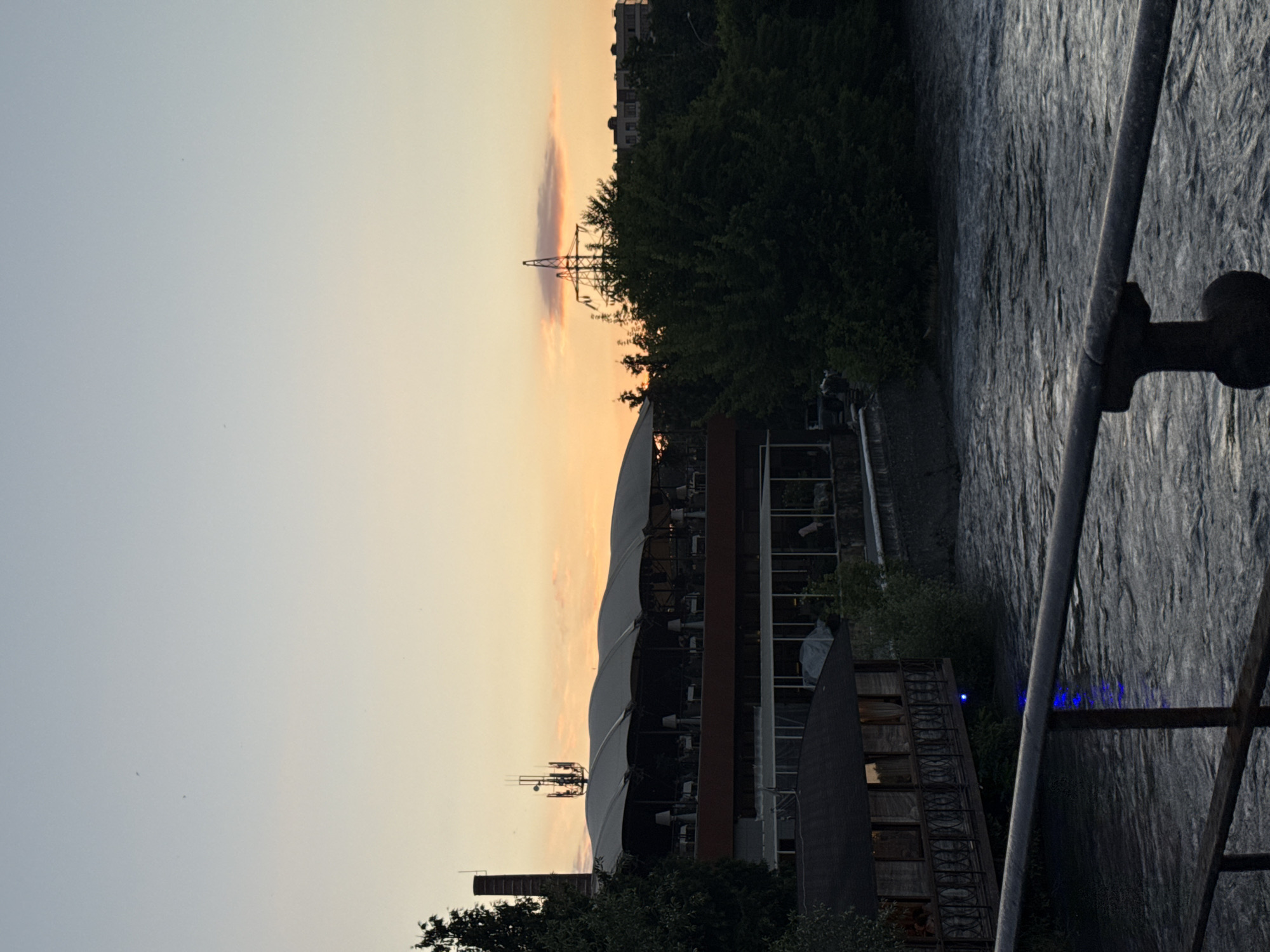 This vertical, eye-level, medium shot depicts a riverfront scene at dusk, likely in Astrakhan, Russia. The sky is a gradient of pale blue transitioning to warm orange near the horizon. In the middle ground, a large, multi-story building with a distinctive arched, covered roof extends across the frame. This building appears to be a restaurant or entertainment venue, with tables and chairs visible on its upper level. A tall, brick smokestack stands to the left of this structure. To the right of the main building, a dense cluster of dark green trees is prominent. A lattice-like electricity pylon stands amidst the trees, silhouetted against the colorful sky. In the foreground, a dark metal railing runs horizontally across the lower portion of the image, obscuring a portion of the river. The river itself is a turbulent, dark gray, with visible ripples and currents. A small, dark wooden structure with windows and a railing, possibly a covered walkway or a small building, juts out over the river on the left side, with a faint blue light visible beneath it. Buildings and trees are also visible on the far bank of the river, appearing as dark shapes against the twilight sky. No people are clearly identifiable in the image. The overall impression is one of a relaxed, evening ambiance, with the natural elements of the river and sky contrasting with the man-made structures of the city.