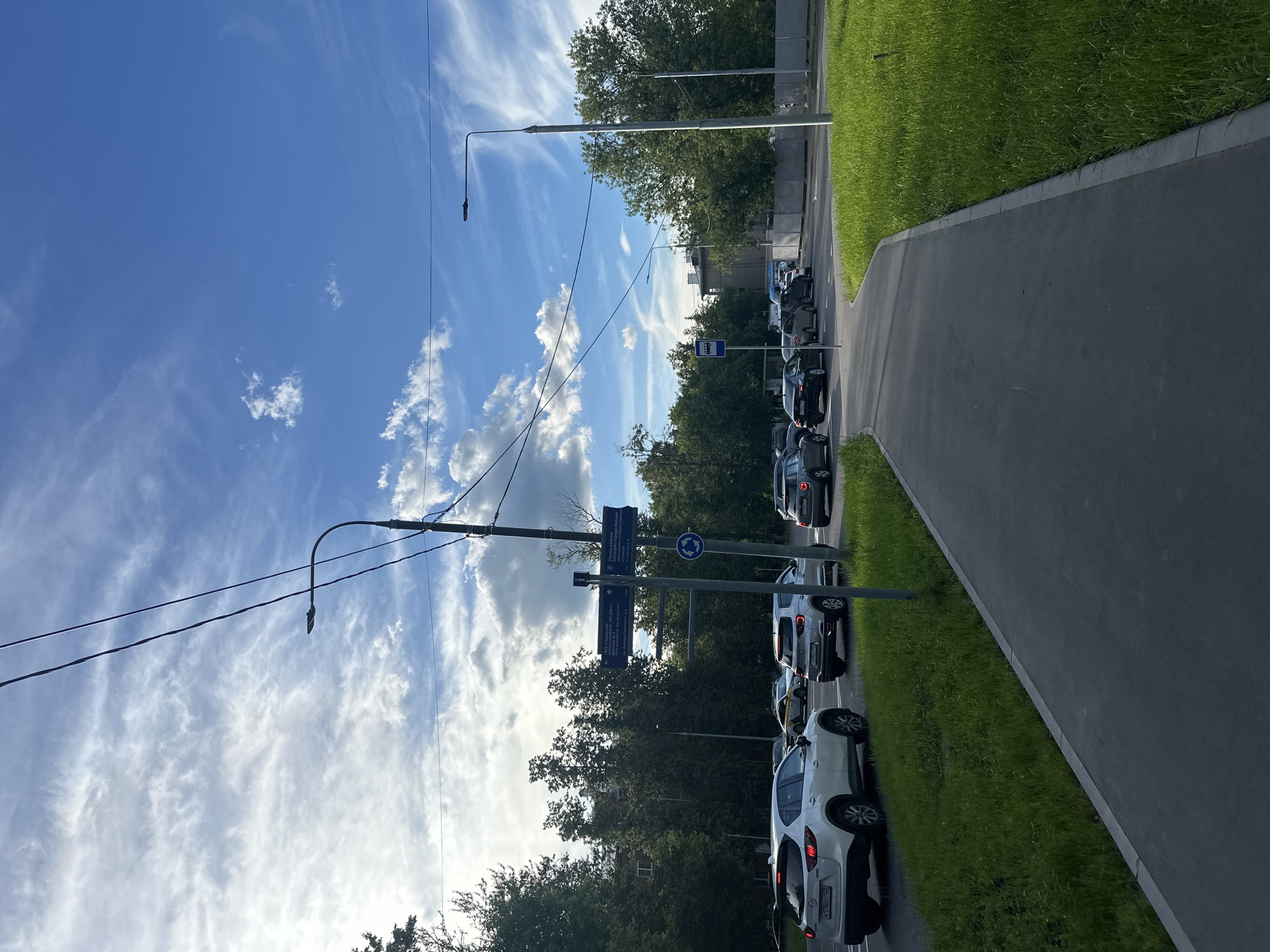 The image depicts a street scene in Moscow, Russia, on a bright day with scattered clouds. A paved walkway runs along the right side of the frame, bordered by a grassy area. A road with several cars in traffic is visible to the left of the walkway. The cars are mostly SUVs and sedans, with some in motion and others stopped.
Several streetlights and utility poles with overhead wires are present. A blue directional signpost stands near the intersection, indicating various destinations with Russian text and arrows. One sign has text that appears to read "Управа района" (District Administration). A blue bus stop sign is also visible. In the background, there are trees and buildings. The overall atmosphere suggests an urban environment with moderate traffic.
