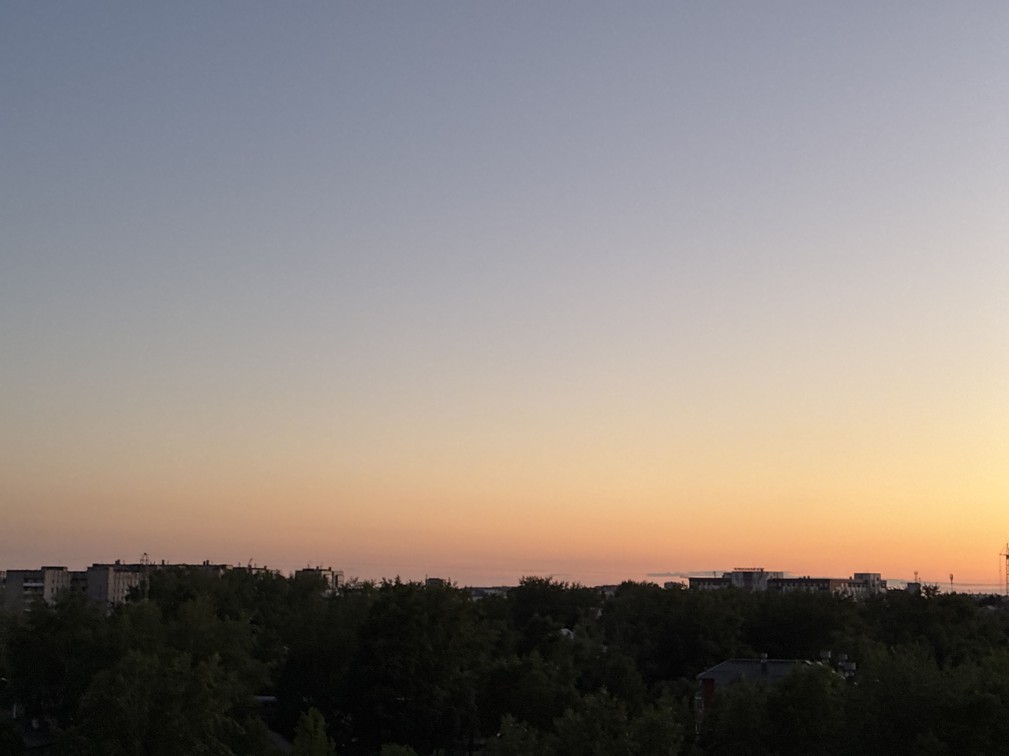 The image captures a serene city skyline at dusk. The sky transitions from a pale blue at the top to a warm gradient of orange and yellow near the horizon, indicating the setting sun. Silhouetted against the colorful sky are the tops of dense trees, forming a dark, lush canopy across the lower portion of the frame. Behind the trees, the outlines of several multi-story buildings are visible, suggesting an urban environment. The architecture of these buildings appears to be consistent with residential or apartment complexes. The overall scene is calm and peaceful, with no people or active events depicted. The location is noted as Unknown, Unknown.