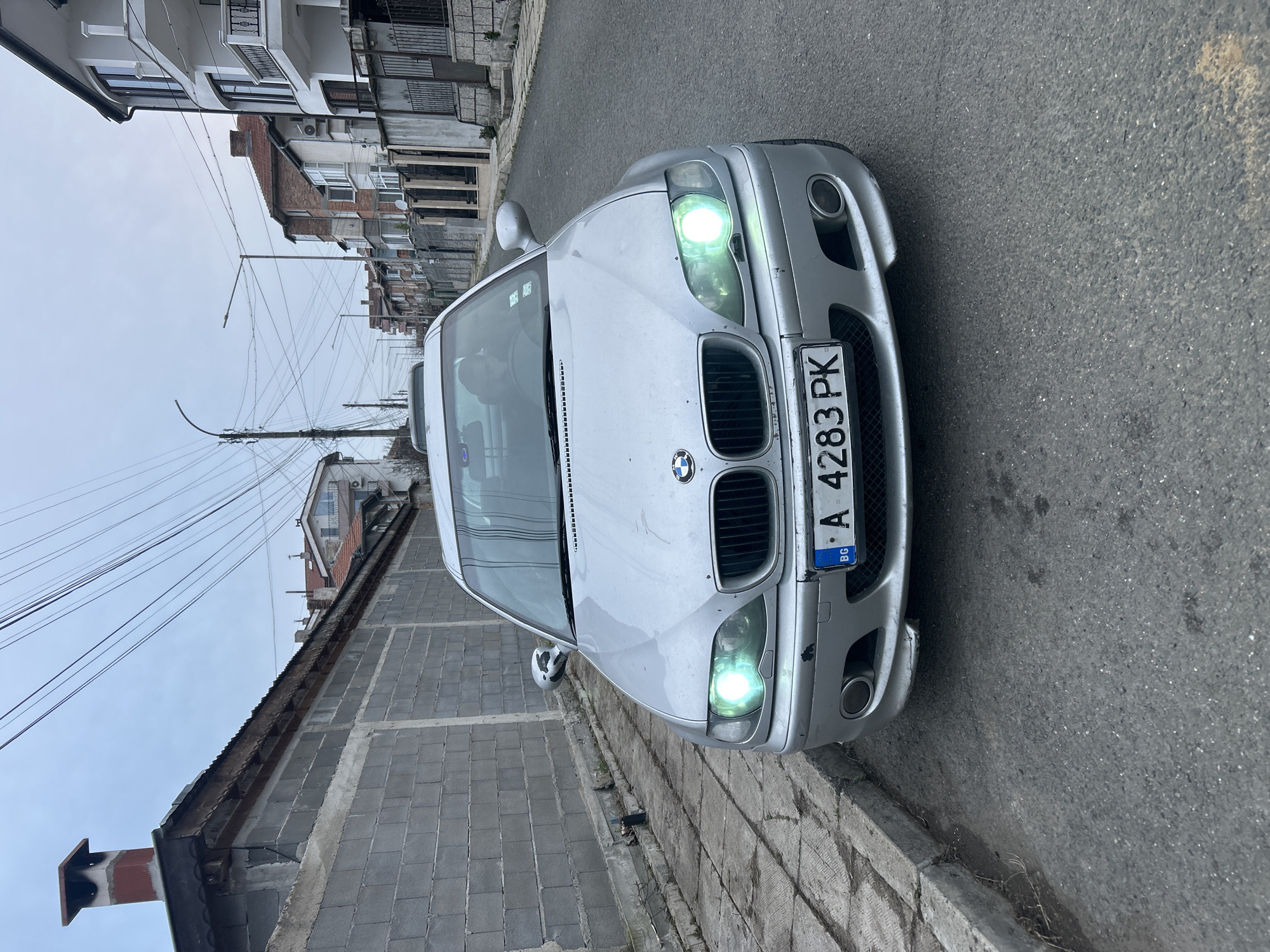 The image shows a silver BMW car parked on the side of a street. The car has the Bulgarian license plate "A 4283 PK". The headlights are on. The background consists of buildings and power lines against an overcast sky. The street appears to be in a residential area. The city is Burgas, Bulgaria.