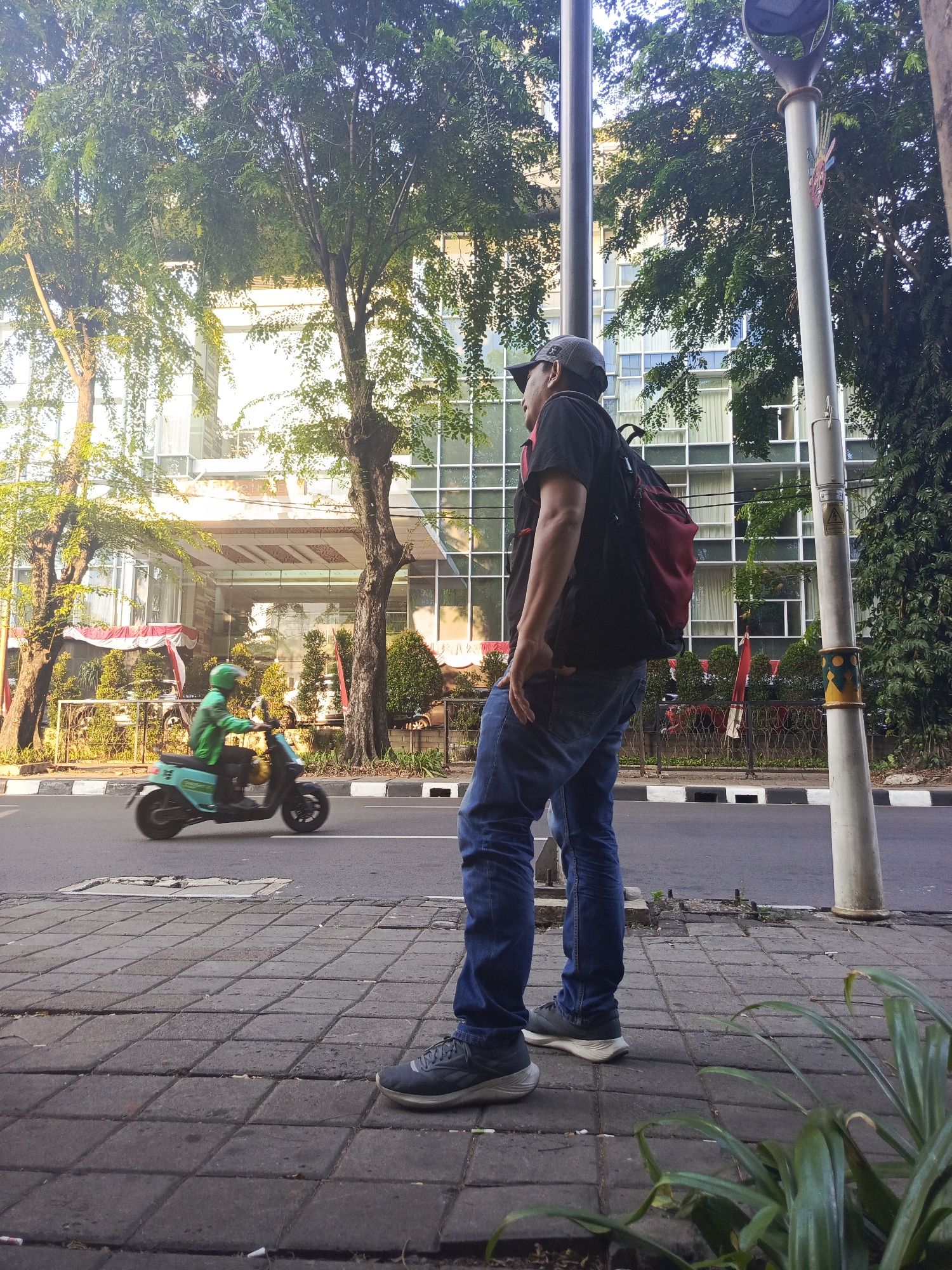 A man wearing a black t-shirt, blue jeans, a grey baseball cap, and a red and black backpack stands on a paved sidewalk. He is looking to the left, with his right hand in his jeans pocket. Next to him, a lamppost is visible. Across the street, a person wearing a green jacket and helmet is riding a teal colored scooter. In the background, a modern building with a glass facade and trees line the street. The scene appears to be in a city.