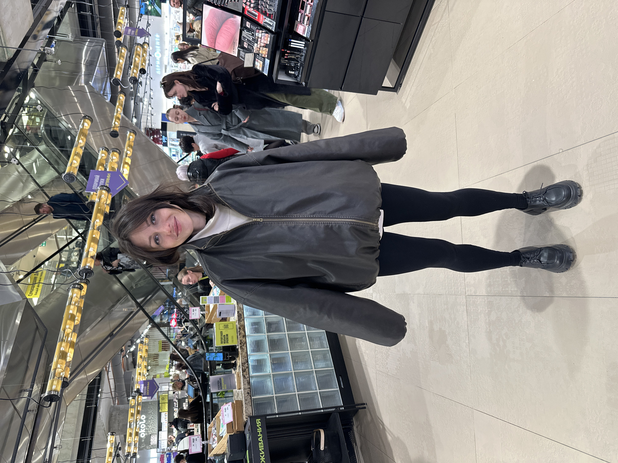 A full-body indoor shot of a woman wearing black leggings, black sneakers, and a dark gray jacket. She is in a shopping mall-type environment; an escalator is visible in the background. The background shows a brightly lit area with various display cases of merchandise and several other people. The image appears to be taken at the Au Pont Rouge department store in Saint Petersburg, Russia.