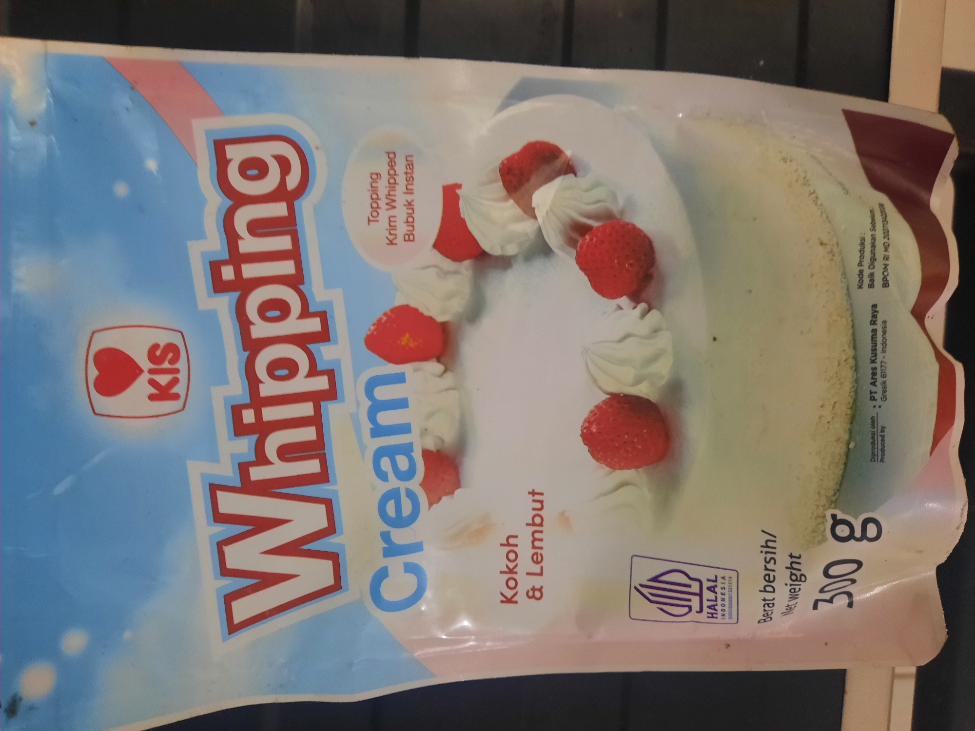 The image is a product shot of a bag of KIS Whipping Cream. The bag is primarily light blue with red and white text. The text "Whipping Cream" is prominently displayed in large, bold, red and white letters. Below that, in blue letters, is the word "Cream". The bag also features an image of a cake topped with whipped cream and strawberries. There is a HALAL logo indicating it is Halal Indonesia. Text at the bottom of the bag provides net weight information as "300 g". Other text indicates production details and expiration information. The product appears to be a food item for baking or dessert preparation. The image itself is a close-up of the product packaging and does not contain any information about Lhokseumawe, Indonesia.