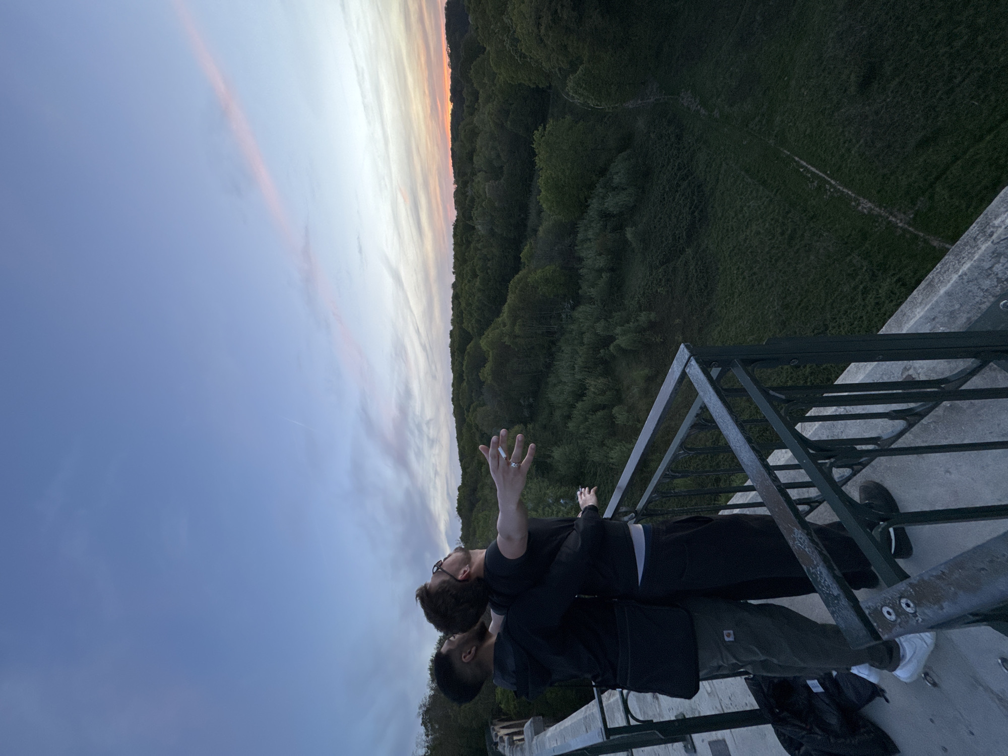 The image depicts two men sharing an intimate moment on a high viewpoint, gazing out over a forested landscape under a twilight sky.
In the foreground, one man with dark skin, short dark hair, wearing a dark top and olive green pants, stands with his back partially to the camera, looking upwards. His left arm is down, and he wears white sneakers. Leaning against him, with his head resting on the first man's shoulder, is a second man with lighter skin, a beard, and glasses. He wears a black t-shirt and black pants. His arms are outstretched wide, palms facing upwards towards the sky, with a lit cigarette held between the fingers of his right hand. A ring is visible on his left ring finger. Both men appear to be absorbed in the vast view and the sky above. A black bag is visible near the first man's feet.
They are positioned behind a dark, somewhat weathered metal railing, possibly part of a bridge, viaduct, or observation deck, which frames the lower portion of the scene. Below the viewpoint, a dense, dark green forest stretches across rolling terrain, fading into the distance. Beyond the tree line, on a sloping expanse of grass, faint, winding paths are discernible.
The sky, dominating the upper part of the image, displays the beautiful hues of dusk or dawn. It transitions from a soft blue on the left to gentle pinks, oranges, and pale yellows towards the horizon, indicating the sun has either just set or is about to rise. The overall atmosphere conveys a sense of peace, freedom, and shared wonder. No visible text is present in the image.