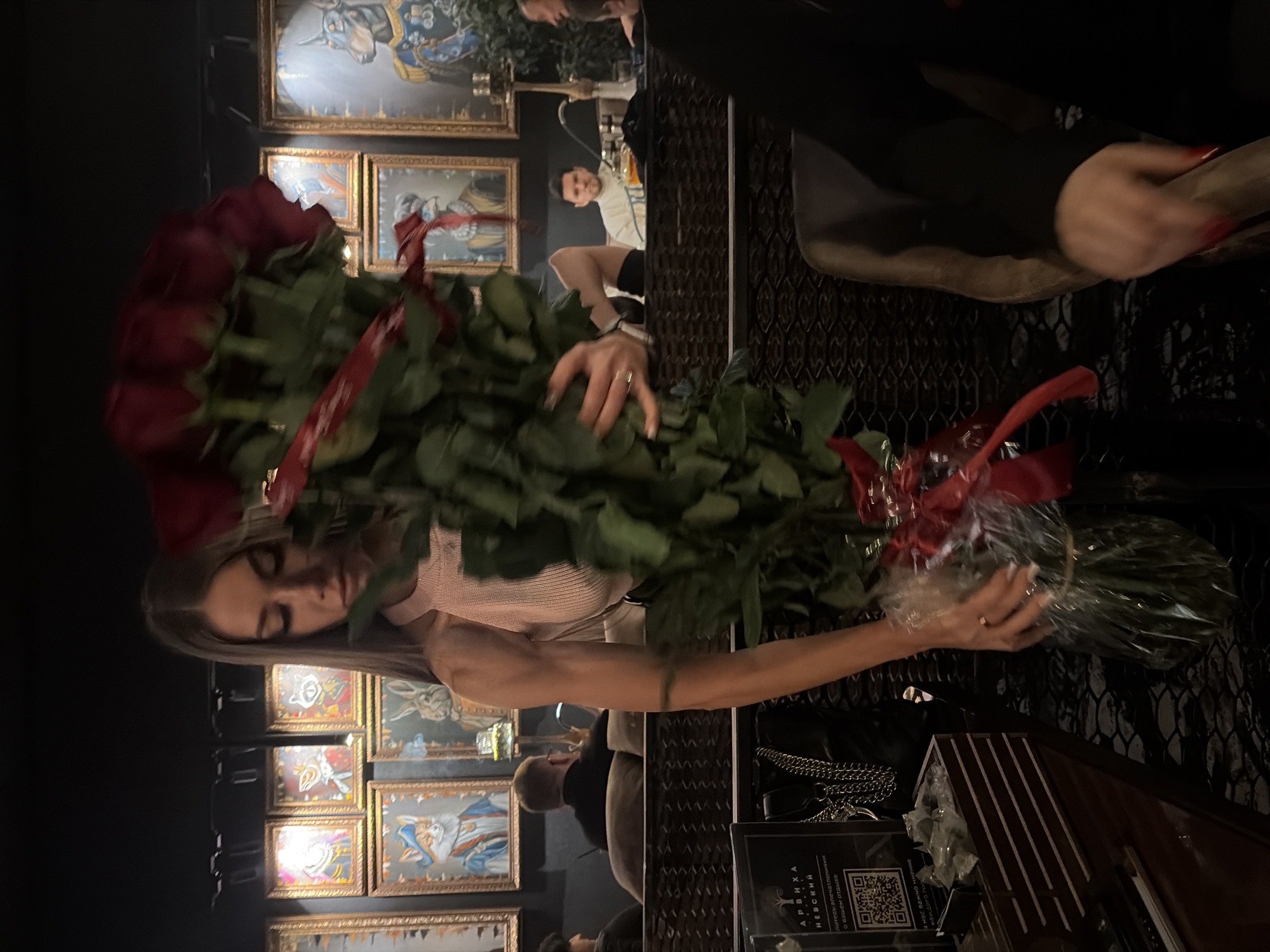 Here is a description of the image:
The image shows a woman holding a large bouquet of red roses in what appears to be an indoor setting, possibly a restaurant or bar. The location is suggested to be in Saint Petersburg, Russia, due to the text on a sign in the foreground mentioning "Арвиха Невский," referencing Nevsky Prospekt, a main street in Saint Petersburg. The woman is wearing a light pink top and has long hair. There are framed pictures on the wall in the background. Other people are seated at tables, and a black handbag is visible on the table next to the woman. The scene appears to be dimly lit.