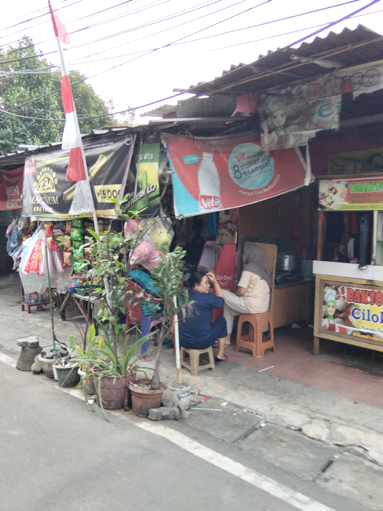 The image depicts a street scene in South Tangerang, Indonesia, likely outside a small shop or food stall. In the foreground, a paved road with a white line marking the edge is visible, with several potted plants lined up along the sidewalk. The plants vary in size and are in different types of pots, some appearing to be made of clay or plastic.
Behind the plants, a store or stall is set up, partially covered by a corrugated metal roof. A red and white Indonesian flag is prominently displayed on a pole extending from the stall. Several banners advertising various products are hung on the front of the stall, including one for "MAGNUM KRETEK" and another with text in Indonesian that appears to relate to mineral water. Bags of goods and merchandise are visible on shelves or hanging from the stall.
Two women are present. One woman, wearing a dark blue outfit, is seated on a low stool and appears to be styling the hair of another woman who is sitting on a small, orange plastic stool. This second woman is wearing a beige headscarf and a light-colored outfit. The background shows the interior of the stall, with some cooking equipment like a pot visible. A sign for "BAKSO CILOK" is displayed on a display case to the right, indicating that food is being sold. The overall scene suggests a bustling local marketplace or a small business operating on the street.