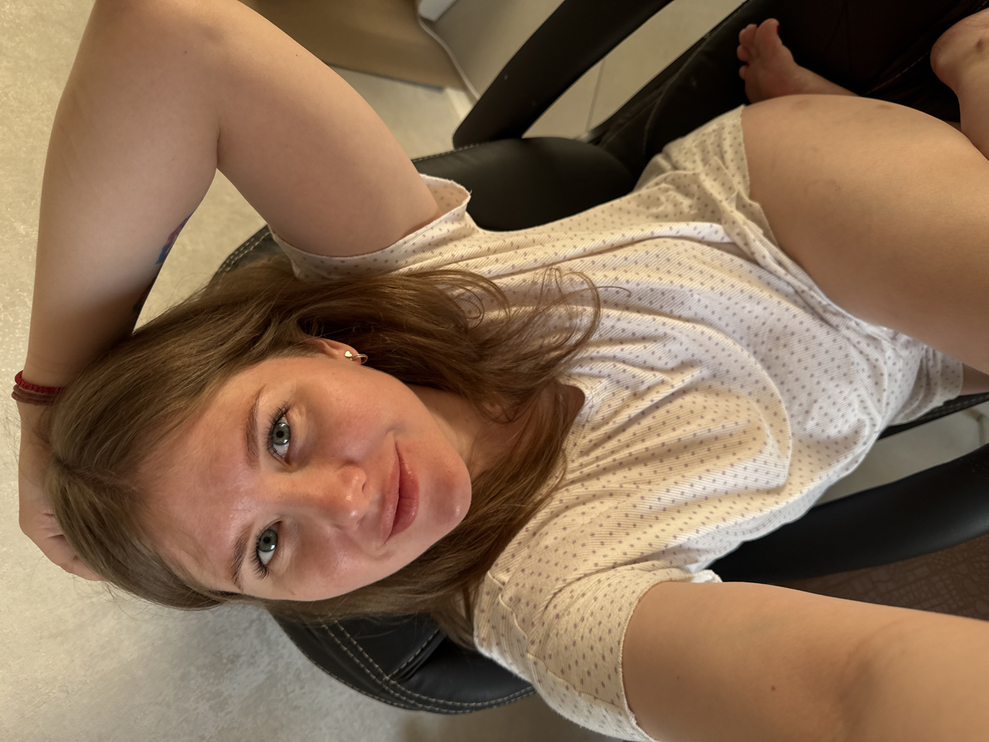 An overhead selfie captures a young woman with light brown hair and striking blue-green eyes, lying back or reclining in a black office chair in what appears to be an indoor setting. She has a natural complexion and a subtle, gentle smile, looking directly at the camera. She wears a light-colored, short-sleeved top adorned with small, light purple polka dots.
Her left arm is raised, with a red bracelet visible on her wrist, and a partial, multi-colored tattoo (featuring blues and possibly pink/red) peeks out from under her sleeve on her upper arm. A small, heart-shaped gold earring is visible in her right ear. Her bare feet are visible in the background, resting on a dark surface, likely part of the chair or an adjacent piece of furniture. The floor around her is a light color, possibly tiled. The overall scene suggests a casual and relaxed moment, possibly at home in Ферма, Russia, during the daytime. There is no visible text in the image.