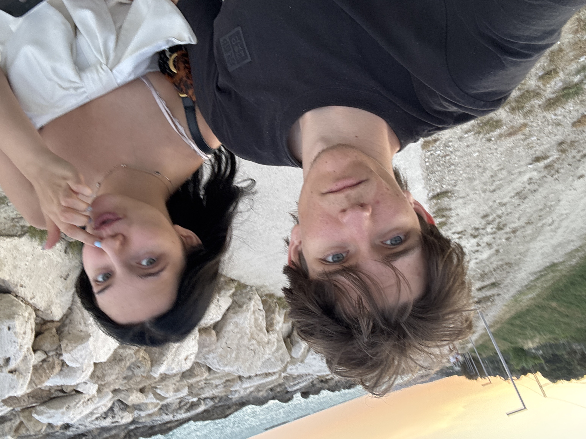 The image shows a selfie of two young adults, a male and a female, standing outdoors near a rocky coastline. The male, in the foreground, has brown hair and is wearing a black t-shirt. The female, to his right, has long dark hair and is wearing a white top. She is touching her nose with her finger, which has light blue nail polish. In the background, there is a path lined with lampposts, the sea, and a rocky shoreline. The sky suggests it might be sunset or sunrise with an orange hue. The scene appears to be a public walkway or park area. The location is identified as Grottammare, Italy, a coastal town.