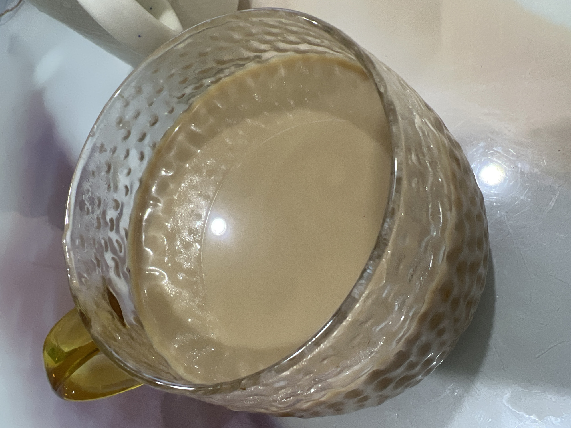 A high-angle, close-up shot depicts a textured glass mug filled with a light-brown liquid, possibly coffee or tea with milk. The mug has a clear body with a yellow handle. The surface of the liquid is reflective, showing a bright spot of light. The mug rests on a white or light-colored surface, potentially a table or countertop. A small portion of a white object, perhaps another mug or container, is visible in the upper-left corner. The photograph was taken indoors. There is no indication of the location being in Chaikovsky, Russia, or any specific geographic location. No people are visible in the image, and no specific actions or interactions are depicted. The focus is solely on the mug and its contents.