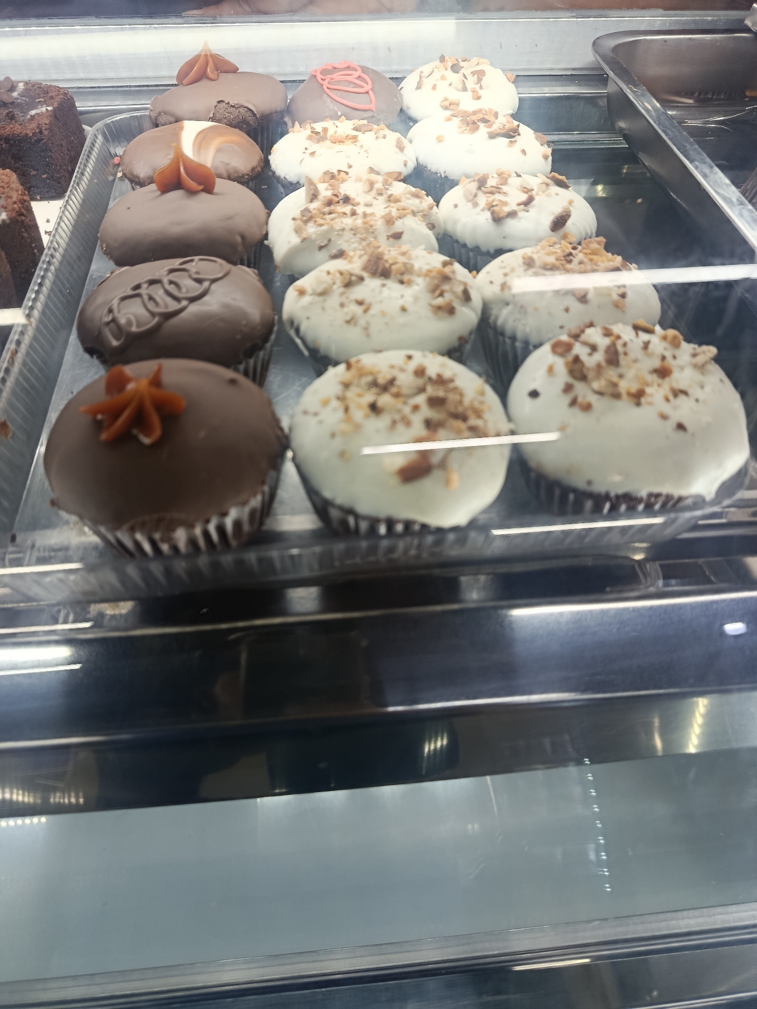 The image shows a display case of cupcakes in Altagracia, Venezuela. The cupcakes are arranged in rows. The front row has chocolate cupcakes with various decorations, including a caramel-colored star, circular designs, and caramel swirls. The second row consists of white frosted cupcakes topped with chopped nuts. Another tray of brownies sits to the left of the cupcakes. The display case is made of metal with a glass front. The focus is on the cupcakes and their arrangements.