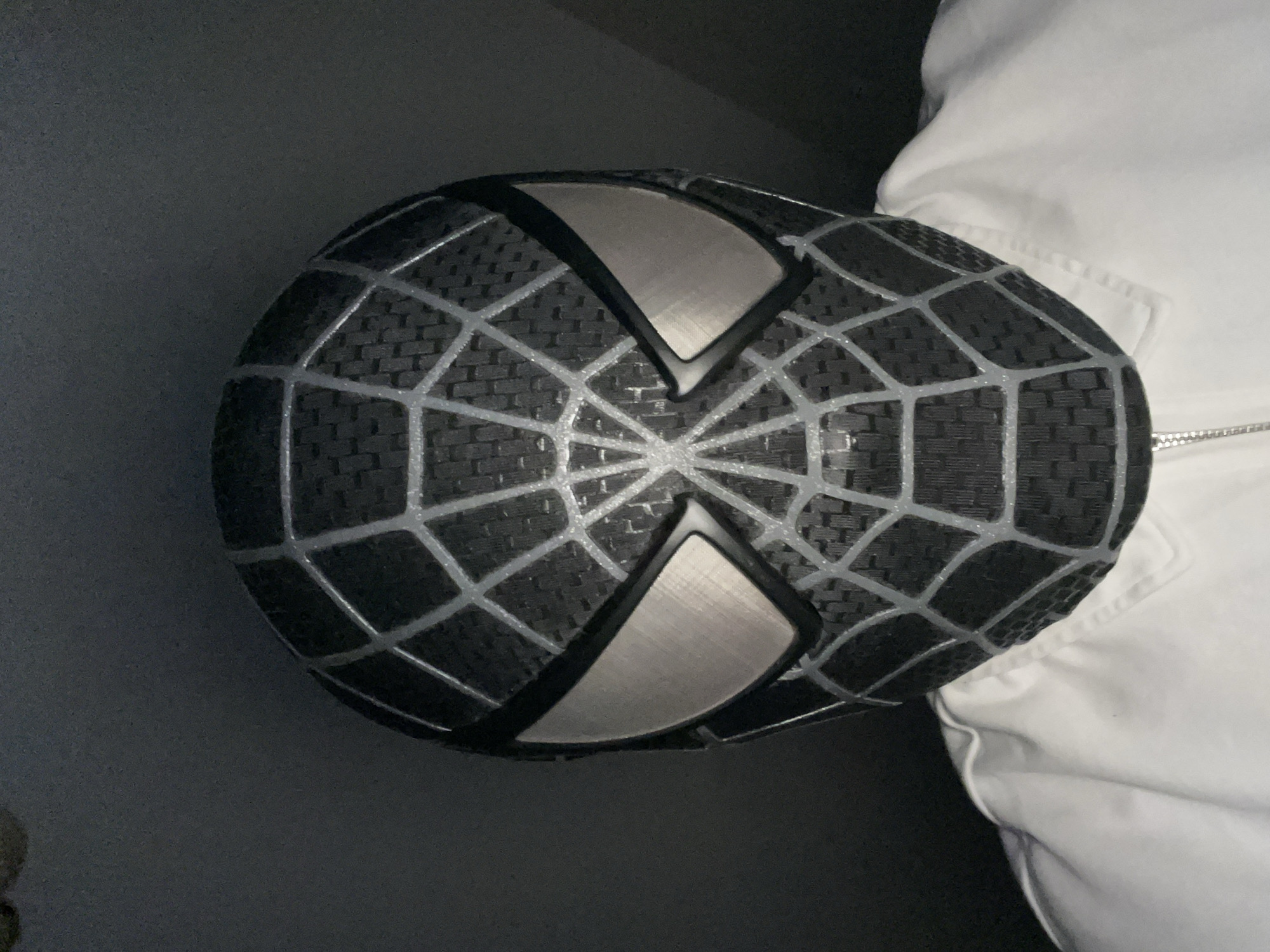 The image displays a close-up, angled view of an object resembling a black Spider-Man mask, specifically the Symbiote suit design. The mask is predominantly black with a distinct woven or textured pattern, similar to carbon fiber or a specialized fabric. Overlaid on the black material are raised silver or light grey web lines that converge towards the center of the face.
The eye lenses are a prominent feature, shaped distinctly with sharp, pointed inner corners. They appear to be made of a reflective, brushed silver or metallic grey material, outlined by a thin black border. The mask is positioned against a dark, featureless background on the left and bottom.
To the right of the mask, a portion of a white garment is visible. This garment, possibly a jacket or shirt, exhibits creases and folds, and a metallic zipper pull is partially seen near the bottom right. There is no visible activity taking place; the mask appears stationary, possibly resting on a surface or worn by an unseen individual. No text is visible in the image. The lighting is subdued, suggesting an indoor setting or low light conditions. No specific location or time of day cues are discernible from the image itself.