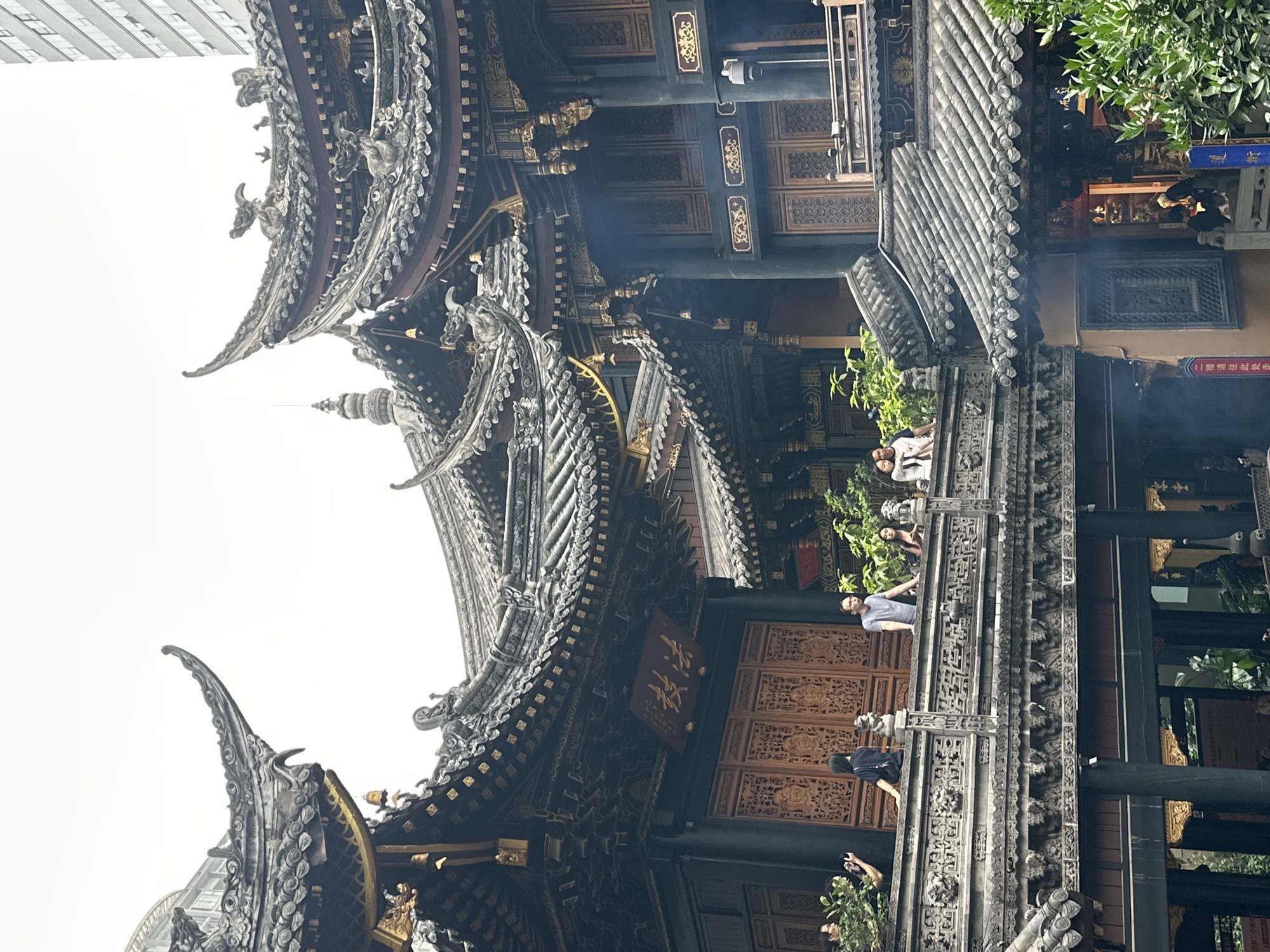 The image depicts a portion of the Arhat Temple in Yuzhong District, Chongqing, China. The traditional Chinese architecture features multi-tiered roofs with elaborate detailing and curved eaves, topped with decorative elements. Several people are visible on a walkway, some interacting with each other and observing the scene. Plants provide a touch of greenery. A modern building appears in the background, contrasting with the ancient temple. Smoke rises from what looks like incense or offerings within the temple grounds.