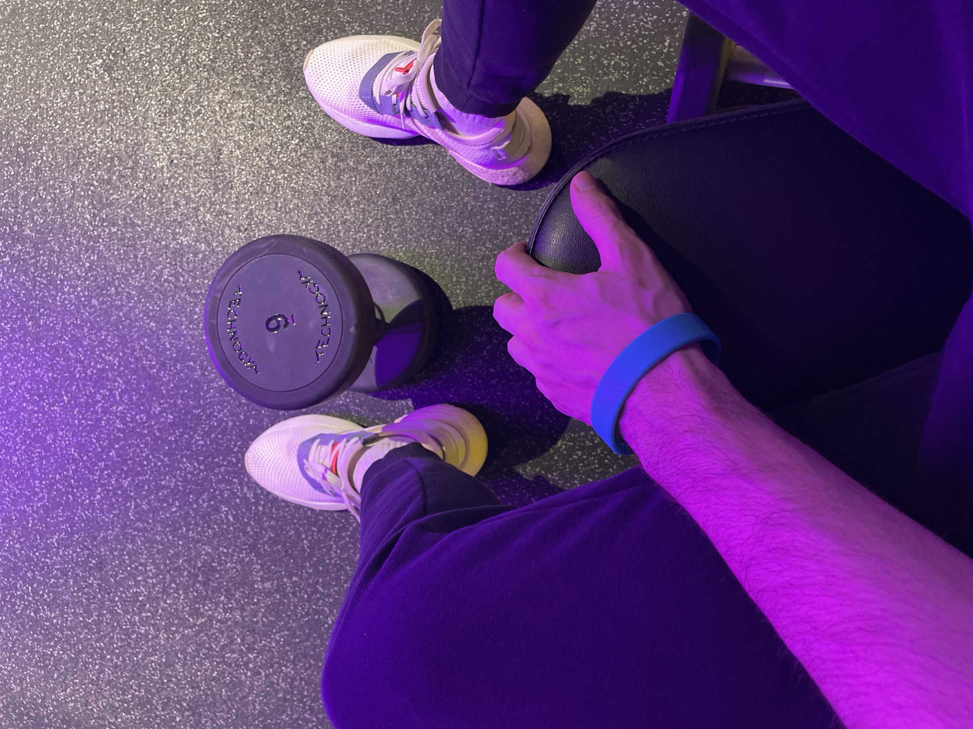 The image depicts a person in a gym setting. The individual is seated on a workout bench, with a 6 kg dumbbell positioned on the floor nearby. The person is wearing athletic shoes, dark sweatpants, and a blue wristband. One hand is visible, resting on the workout bench. The gym floor is visible as well as the bench material. The lighting in the gym appears to be predominantly purple. The image does not contain information to identify any specific location in the city of Saint Petersburg, Russia.