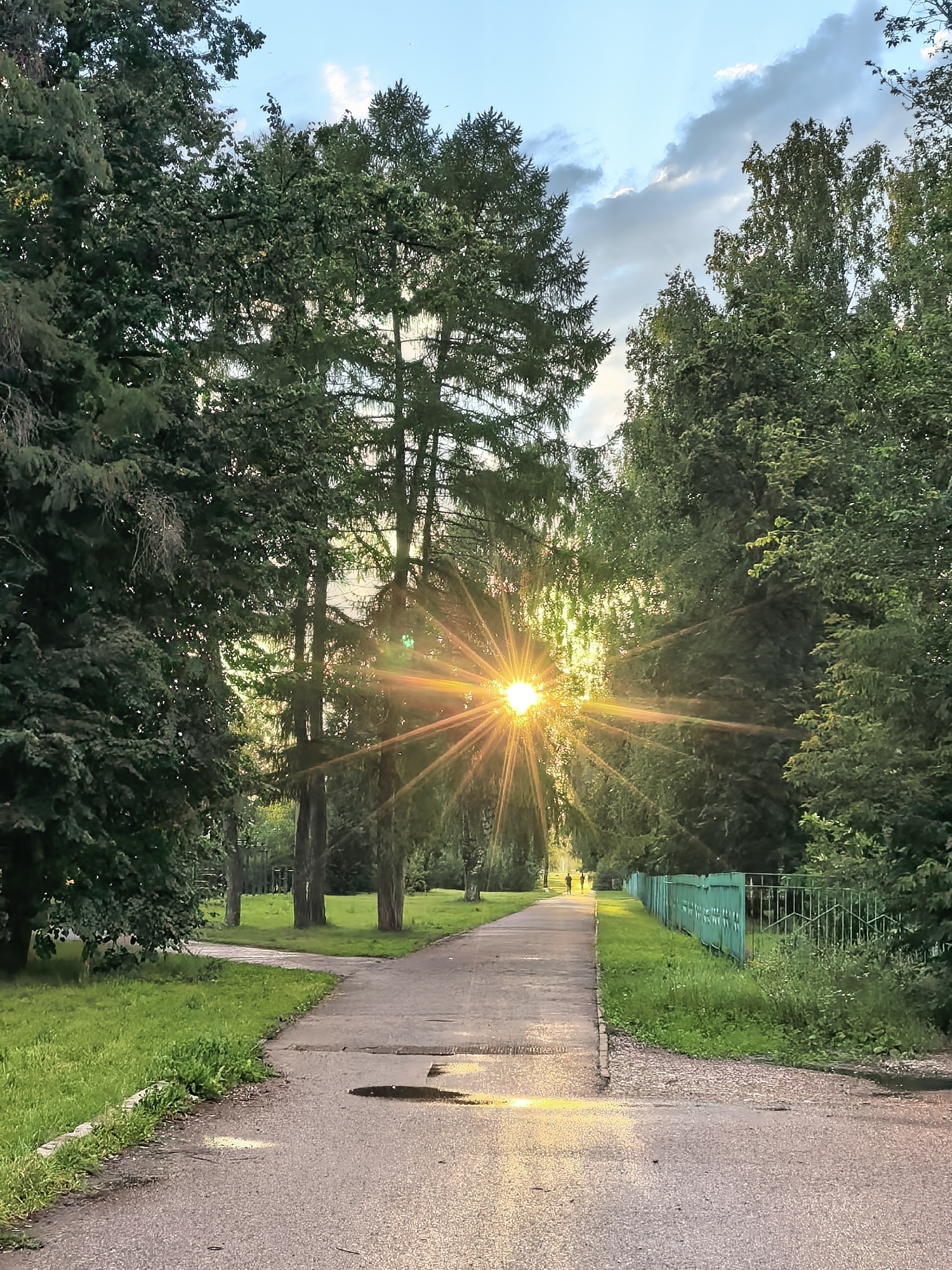 The image depicts a park path in Sterlitamak, Russia, under a partly cloudy sky at sunset. The sun's rays are piercing through tall trees, creating a starburst effect. The path, made of asphalt, shows signs of wear and has puddles from recent rain. Lush green grass borders the path, and a light green metal fence runs along the right side. Two silhouetted figures can be seen walking in the distance on the path. The overall impression is one of a serene, late afternoon in an urban park.