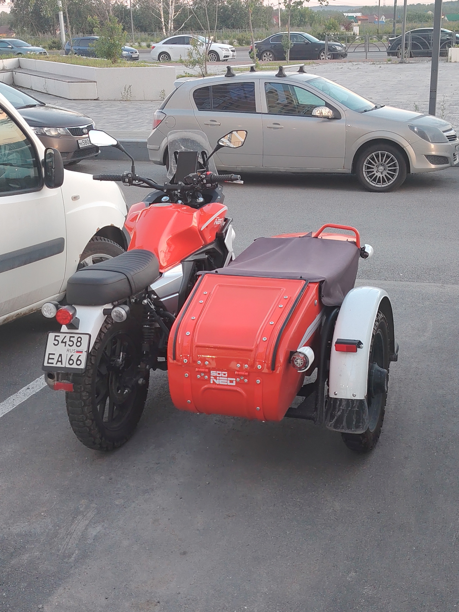 The media file displays a red motorcycle with a sidecar parked on an asphalt surface, likely a parking lot or street. The motorcycle is positioned to the right of a white vehicle. The sidecar is also red and appears to be covered with a dark fabric.
The background shows a line of parked cars and a building in the distance. Trees and foliage are visible, suggesting an outdoor setting, possibly during daytime, given the lighting. The overall scene appears to be a casual, everyday moment.
Visible text on the motorcycle's license plate reads "5458 RUSS EA 66". The sidecar also has some text printed on it, including "500" and "NEO".
The composition of the image focuses on the motorcycle and sidecar, with the white vehicle partially visible to the left. The perspective is from a slightly elevated viewpoint, looking down at the vehicles.