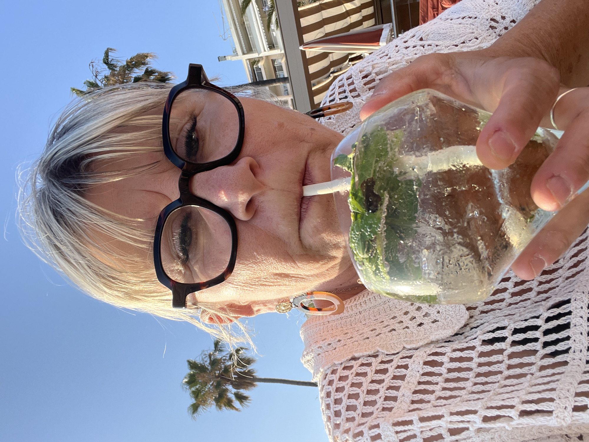 A woman with blonde hair, wearing glasses and a white crochet top, is taking a sip from a glass of iced beverage. She is outdoors, with palm trees and buildings visible in the background. The scene suggests a sunny day, possibly in Cannes, France, given the palm trees and coastal architecture. The woman's expression is neutral as she drinks. The glass contains ice and greenery, likely mint.