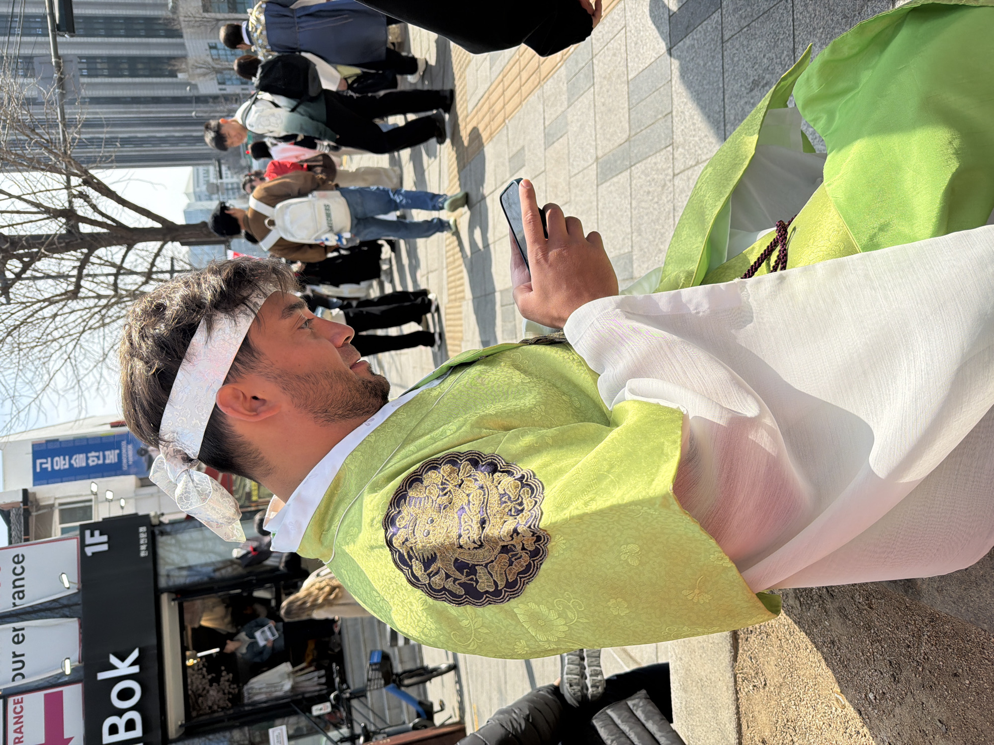 A male individual is seated outdoors on a paved pedestrian area, focused on a black smartphone held in both hands. He is seen in profile, facing right, with dark, curly hair and a beard. He wears a traditional Korean Hanbok, consisting of a vibrant lime-green outer robe (po or jeogori) detailed with a round, embroidered emblem of a mythical creature in dark blue and gold on the left shoulder, and a white undergarment or outer layer. A patterned white headband with a bow is tied around his forehead.
The scene is located in Insadong, Seoul, South Korea, as confirmed by a blue vertical sign in the mid-background displaying "인사동문화의거리" (Insadong Cultural Street) and "INSA-DONG SEOUL." To the left, a storefront with a black facade and partially visible signage reading "Bok," "1F," and Korean text identifying it as a "Traditional Hanbok Clothing" shop is present. A blue bicycle is parked near the shop.
Numerous pedestrians, dressed in both modern and traditional Korean attire, occupy the background, traversing the paved street. A tall, leafless tree is visible in the mid-ground, and modern high-rise buildings rise in the far distance against a clear sky. The area is brightly illuminated by direct sunlight, casting distinct shadows on the ground.