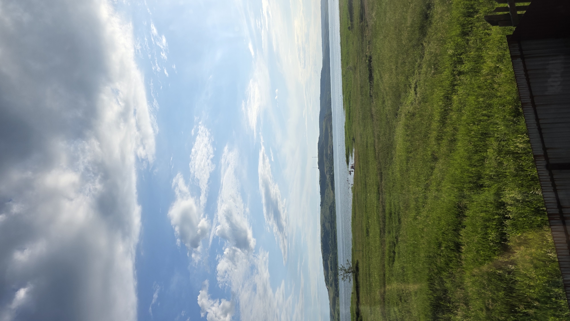 This is a landscape photograph taken outdoors, likely in the early afternoon on a partly cloudy day. The scene depicts a body of water, possibly a river or lake, with green grassy banks on the right side of the frame. In the distance, a treeline or rolling hills are visible along the far bank of the water. A structure, possibly a barn or shed with a corrugated metal roof, is partially visible on the far right.
There are several small figures in the water, some appearing to be on small boats or possibly swimming. The sky is a mix of bright blue with wispy white clouds and some darker, more diffused clouds, suggesting changing weather. The overall mood of the image is serene and natural. There is no visible text within the image.