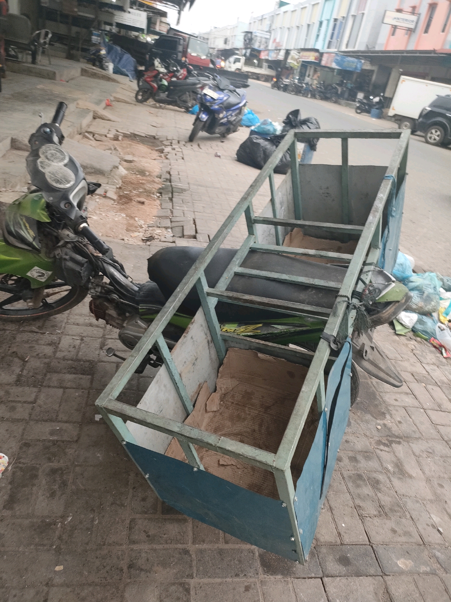 The image shows a close-up view of a motorcycle with a custom-built, metal-framed cargo carrier attached to its side, positioned on a paved sidewalk in Batam, Indonesia. The motorcycle, primarily green with some black accents, is parked facing left, with its front wheel slightly turned. The cargo carrier is a rectangular structure with a metal frame and blue panels on its sides, appearing to be designed for transporting goods. Inside the carrier, there are pieces of cardboard and what appear to be large, dark plastic bags, possibly containing trash or other materials.
In the background, the scene depicts a street with other motorcycles parked, a white truck, and several buildings with awnings and storefronts. The architecture suggests a commercial or market area. The ground is paved with square tiles. There are also several black trash bags and blue plastic bags scattered on the sidewalk near the motorcycle and carrier, indicating a possible refuse collection point or general litter. The overall impression is that of a utilitarian vehicle adapted for transport in a bustling urban environment.