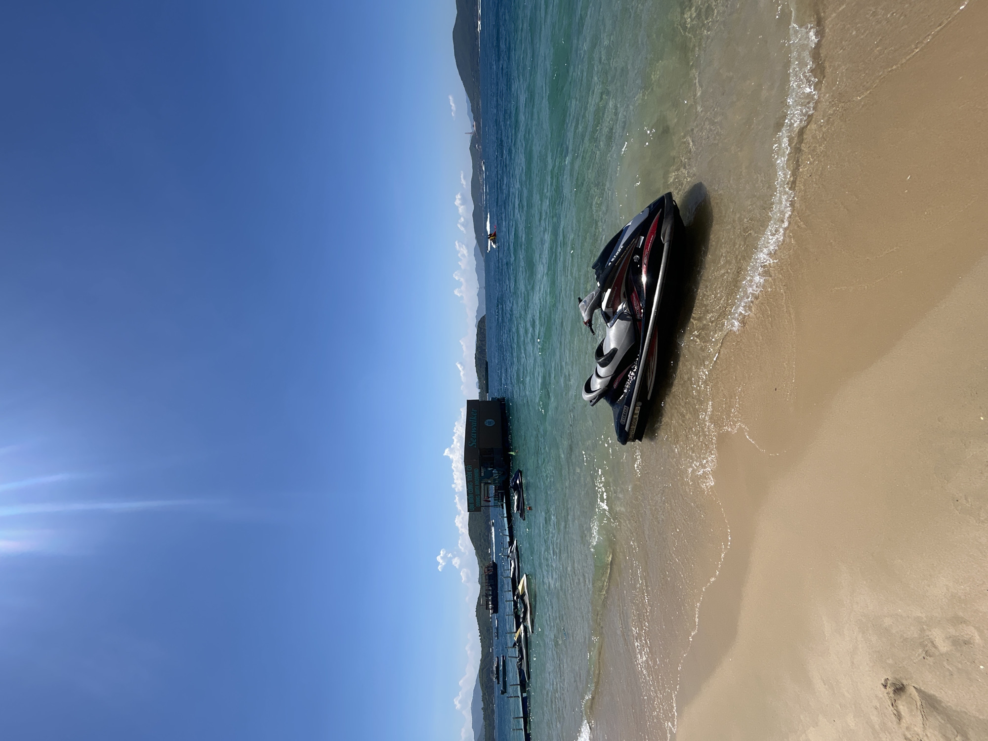 A jet ski is parked on a sandy beach in Bac Nha Trang Ward, Vietnam. The clear blue water laps at the shore where the jet ski rests. In the background, a floating structure with the name "Seawalker" is visible, along with several other jet skis docked at a pier. A larger boat and a few other vessels are scattered across the turquoise water. The sky is a bright, clear blue with a few white clouds, and the sun is shining brightly, creating lens flare. The scene depicts a sunny day at a recreational water activity center.