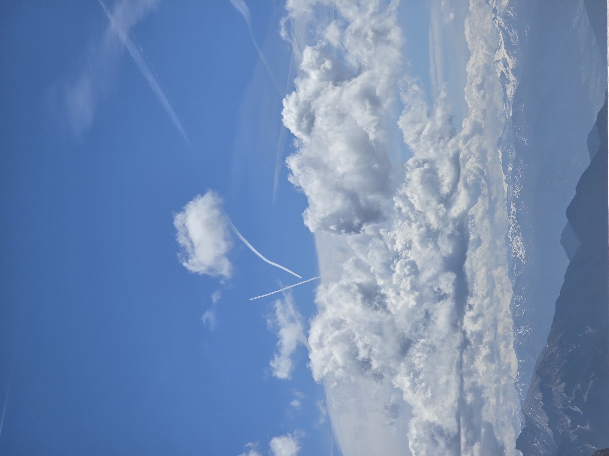 The image depicts a vast, clear blue sky populated with scattered white, fluffy clouds and contrails. In the middle ground, two distinct contrails form a V-shape, suggesting an aircraft's recent passage. Below the sky, a majestic mountain range stretches across the horizon. The peaks of these mountains are partially covered in snow, indicating a high altitude or a cooler season. The foreground is dominated by darker, shadowed mountains, with a hint of what appears to be a body of water at the very bottom edge of the frame. The overall scene is one of natural grandeur, with the sky and mountains dominating the composition. The provided crops confirm the presence of the sky, clouds, contrails, and mountain range. The image is consistent with a view from a high vantage point in or near Garmisch-Partenkirchen, Germany, a region renowned for its alpine scenery and clear skies. There are no people, animals, or man-made structures explicitly visible in the image.