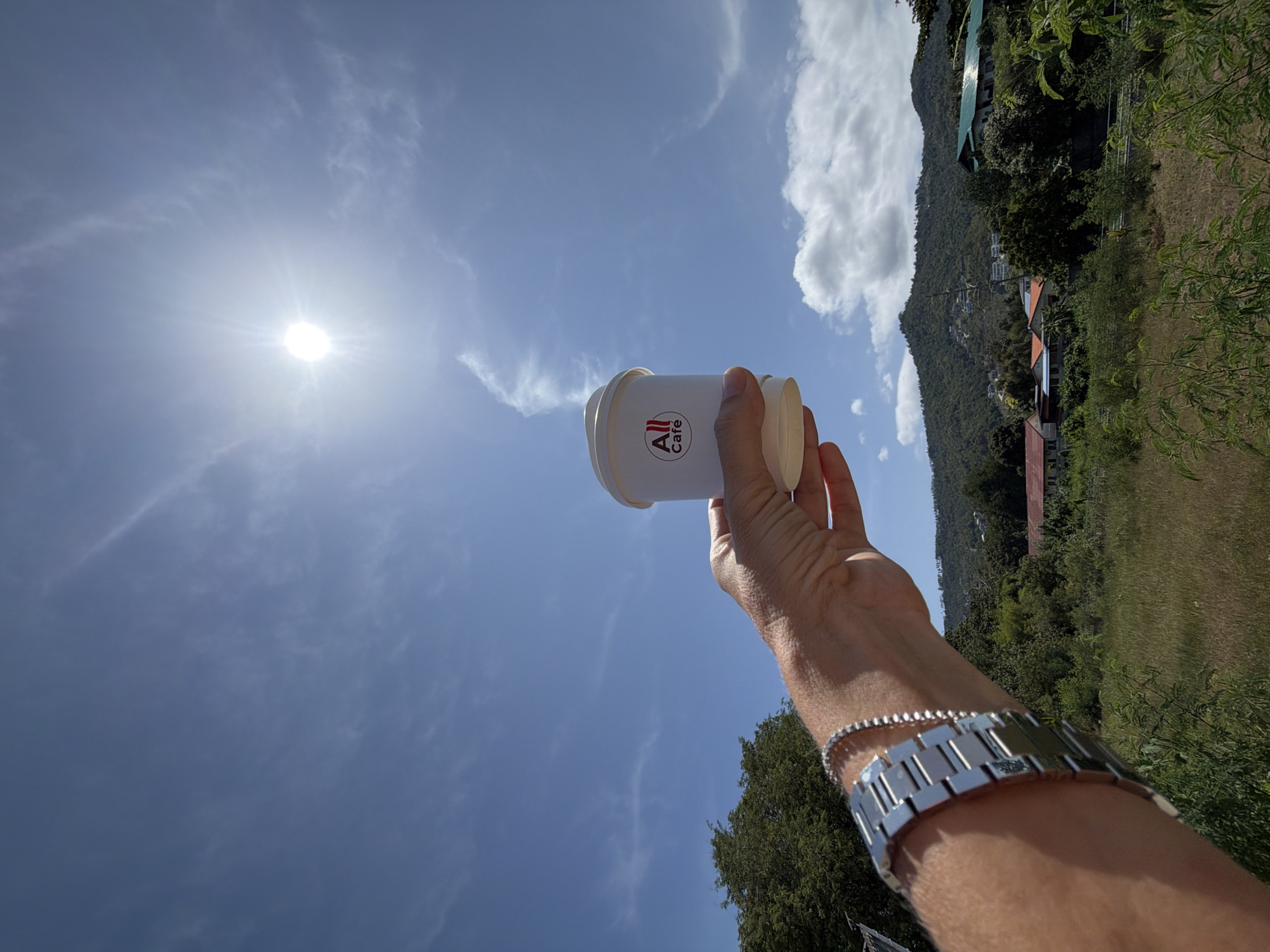 A person's hand, adorned with a silver watch and bracelet, holds a white coffee cup with the "All Café" logo against a bright blue sky with the sun and some clouds. The hand is positioned in the foreground, and the cup is held up towards the sky. In the background, there are lush green trees, hills with houses, and some vegetation. The scene appears to be outdoors, likely in a tropical location like Ko Samui District, Thailand, given the vegetation and sunny weather.