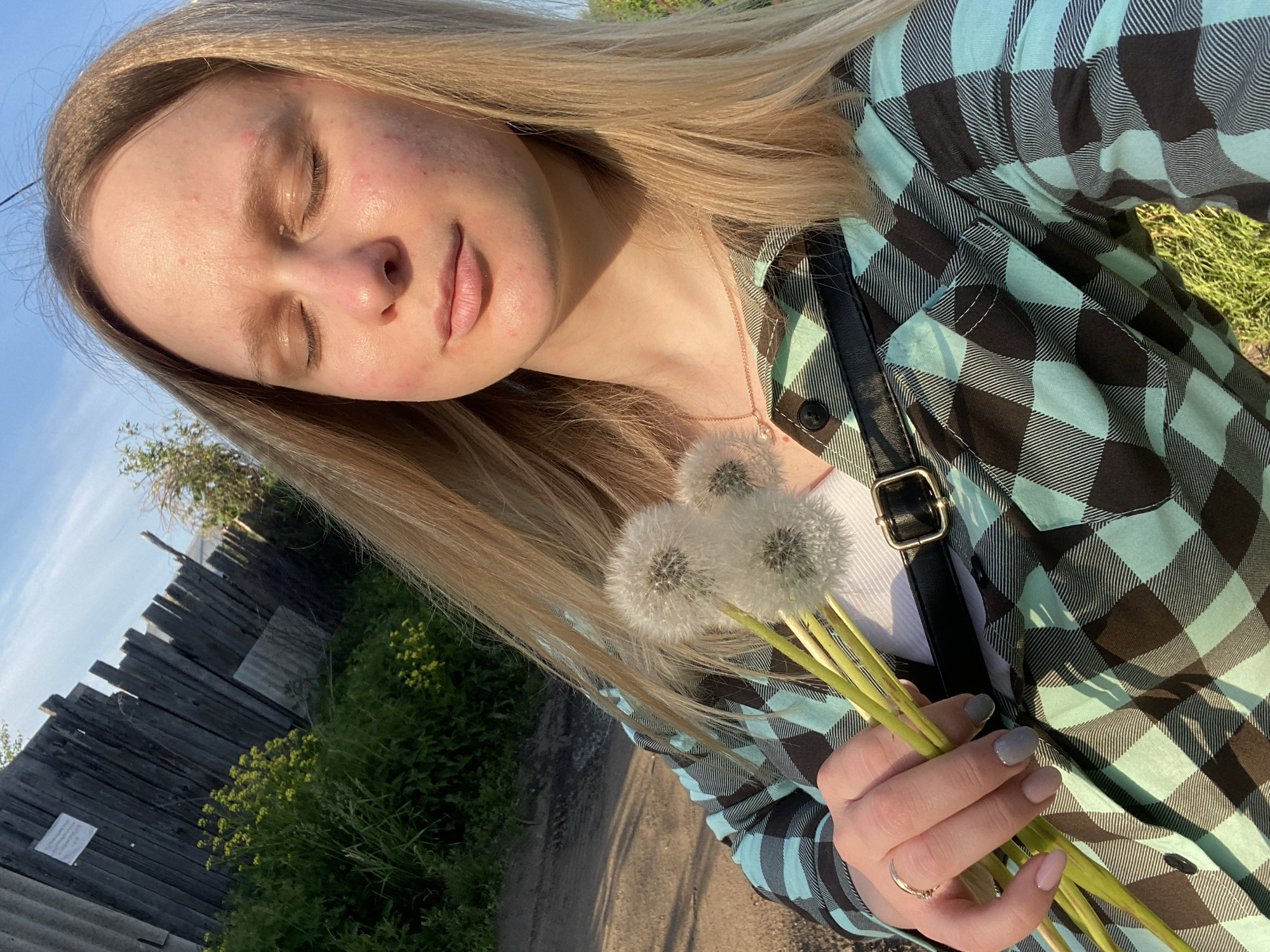 A young woman with long blonde hair is depicted outdoors, bathed in warm sunlight, with her eyes closed and a peaceful expression. Her head is tilted slightly back, suggesting she is enjoying the sun. She holds a small bouquet of several fluffy white dandelion seed heads in her right hand.
She is wearing a plaid shirt featuring light blue (or mint green) and black checks, layered over a white top. A black strap, likely from a bag or camera, is visible over her left shoulder. Her fingernails are painted in shades of light grey and pale pink, and she wears a simple ring on her right ring finger.
The background shows a rustic wooden fence to her left, with lush green foliage and small yellow flowers growing alongside it. A dirt path or road is visible in the lower part of the frame, and a clear blue sky dominates the upper background. The golden quality of the light suggests it is either late afternoon or early morning. A small, white piece of paper is attached to the wooden fence, but any text on it is illegible.
The overall scene conveys a sense of calm and a connection with nature in a rural or semi-rural setting, specifically in Chaikovsky, Russia.